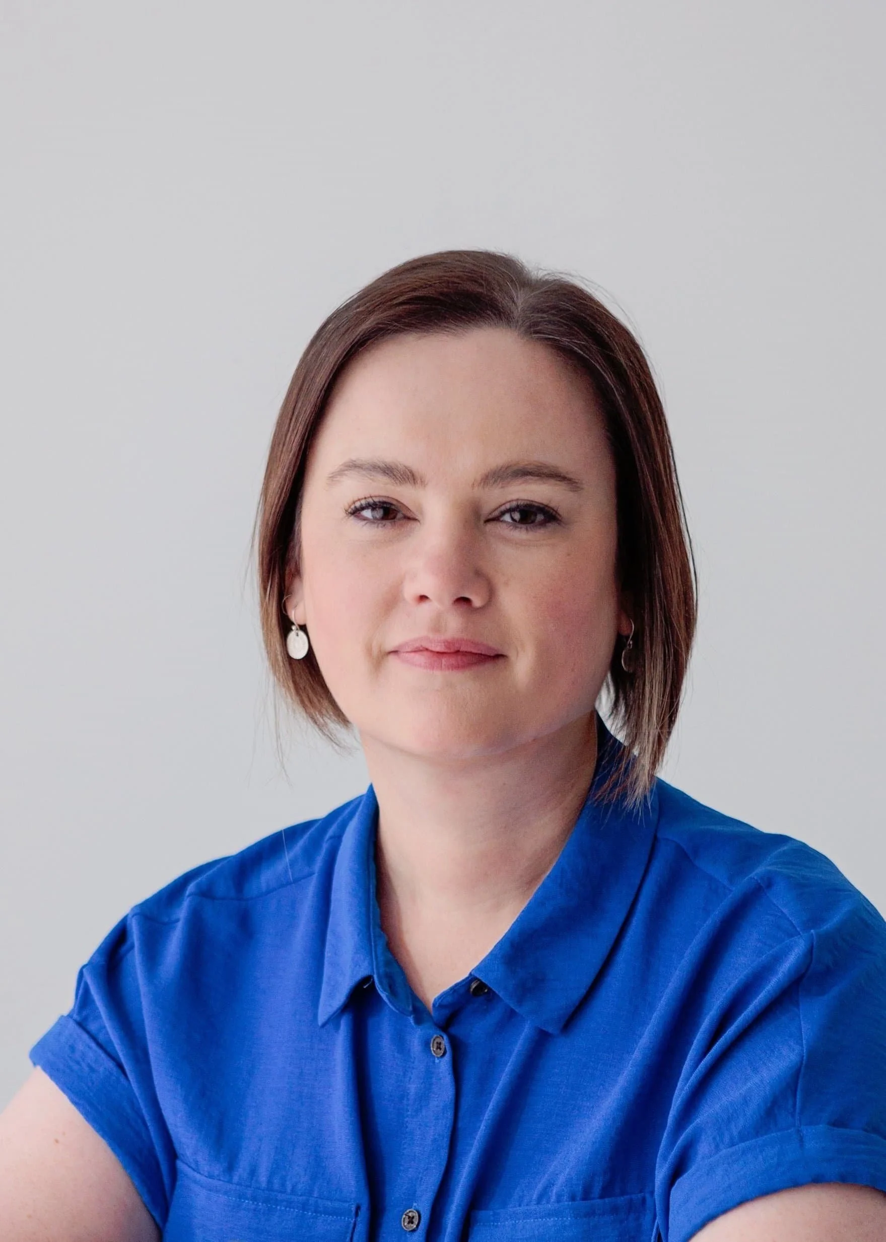 A picture of Louise Hiles. A woman with short brown hair wearing a blue shirt and earrings, looking directly at the camera against a plain white background.