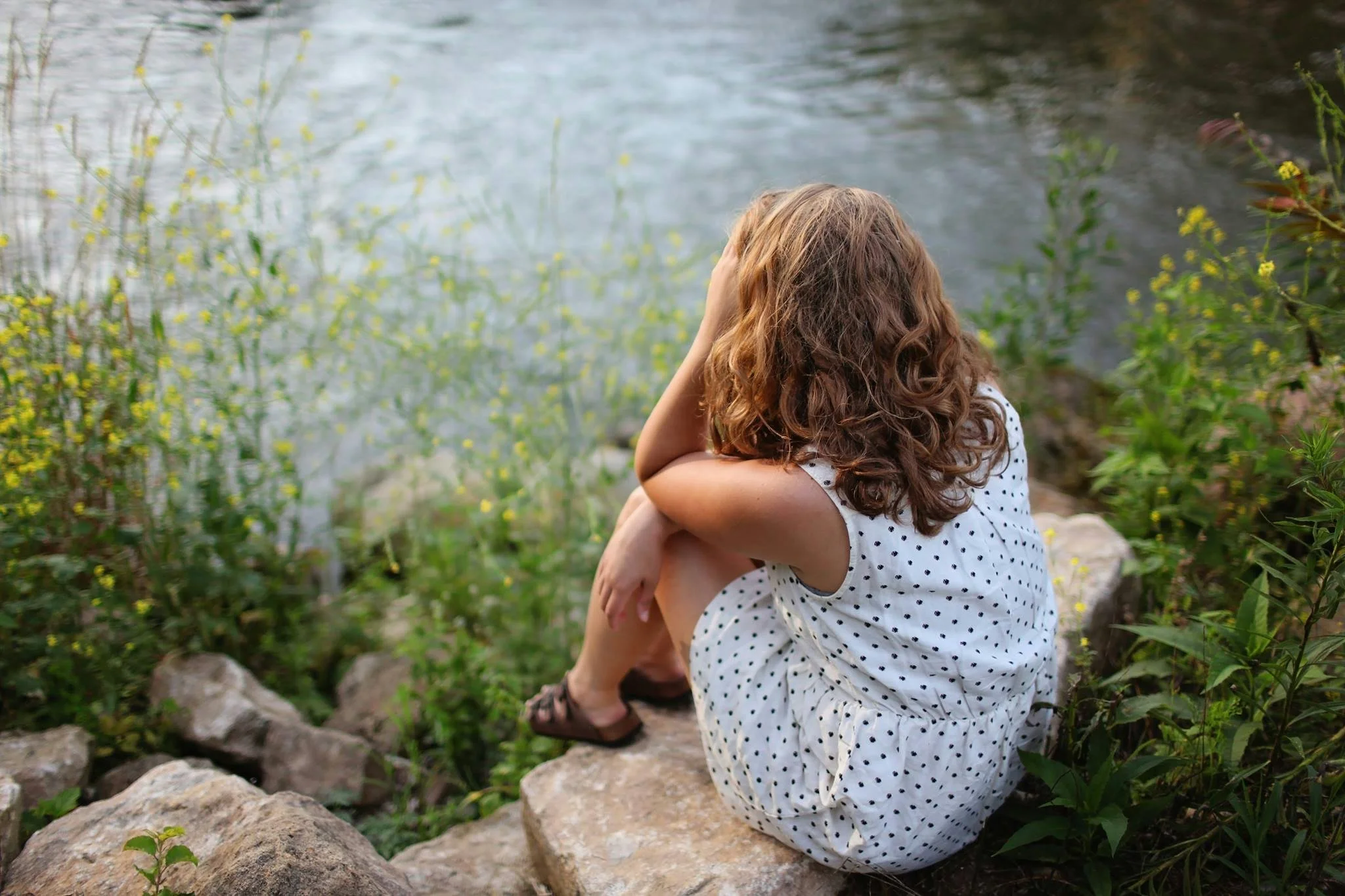 Woman sitting alone by a river, seen from behind, arms wrapped around her knees, surrounded by wildflowers and greenery.