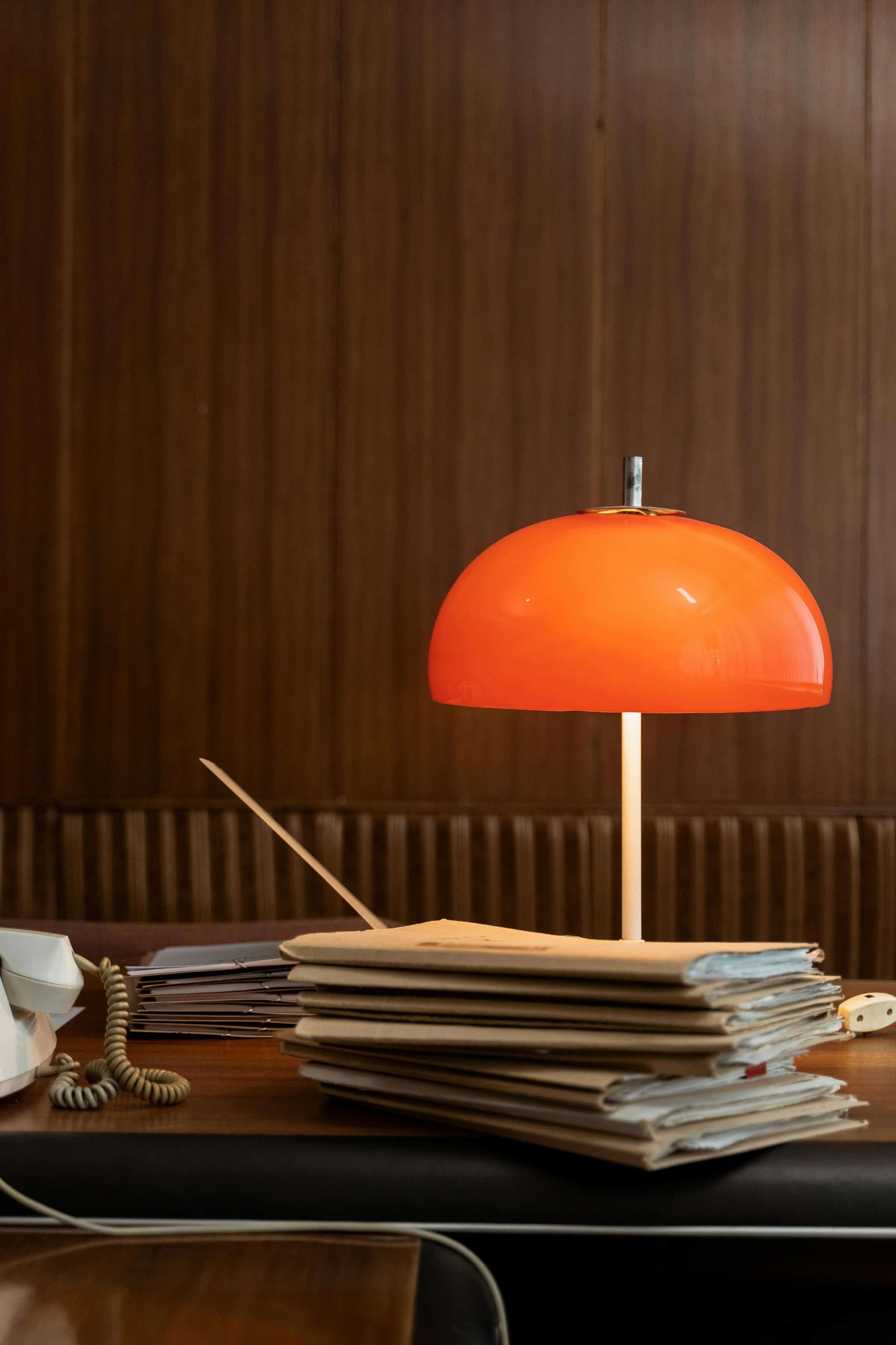 A retro orange desk lamp glows on a wooden desk cluttered with stacked folders and papers, against a wood-paneled wall.