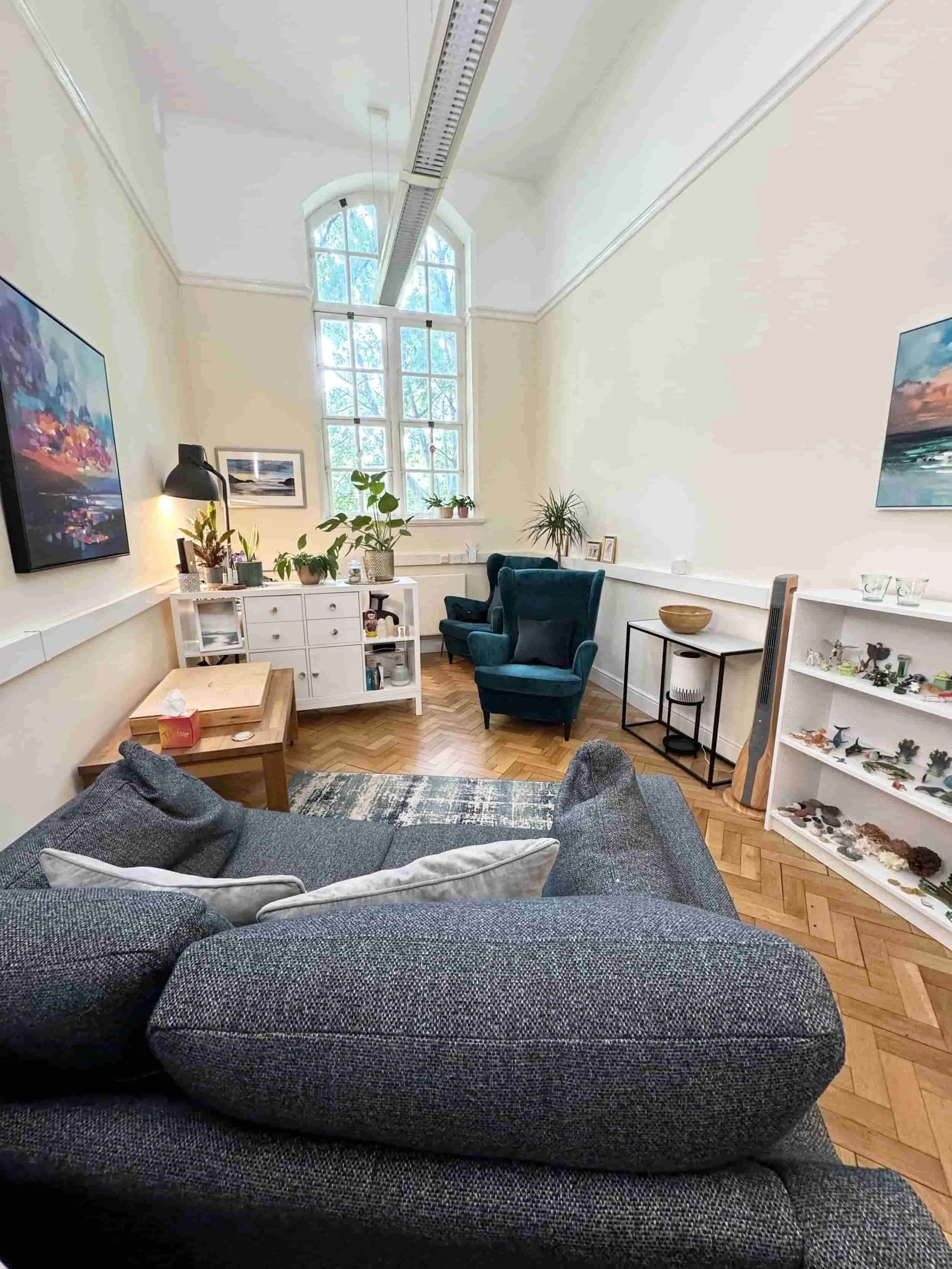 Cozy therapy, coaching consulting room with teal chairs, a sofa, plants, bookshelves with miniatures, art on cream walls, and a large arched window.