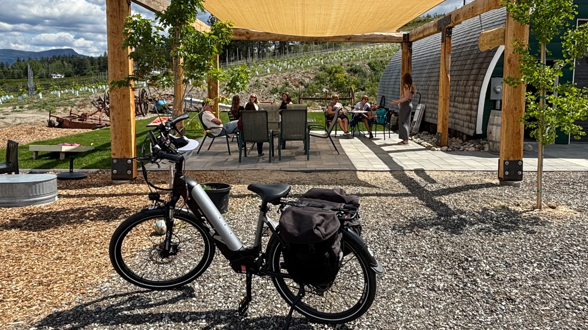 People sitting at an outdoor patio with a wooden pergola, surrounded by trees and hills, with bikes in the foreground.