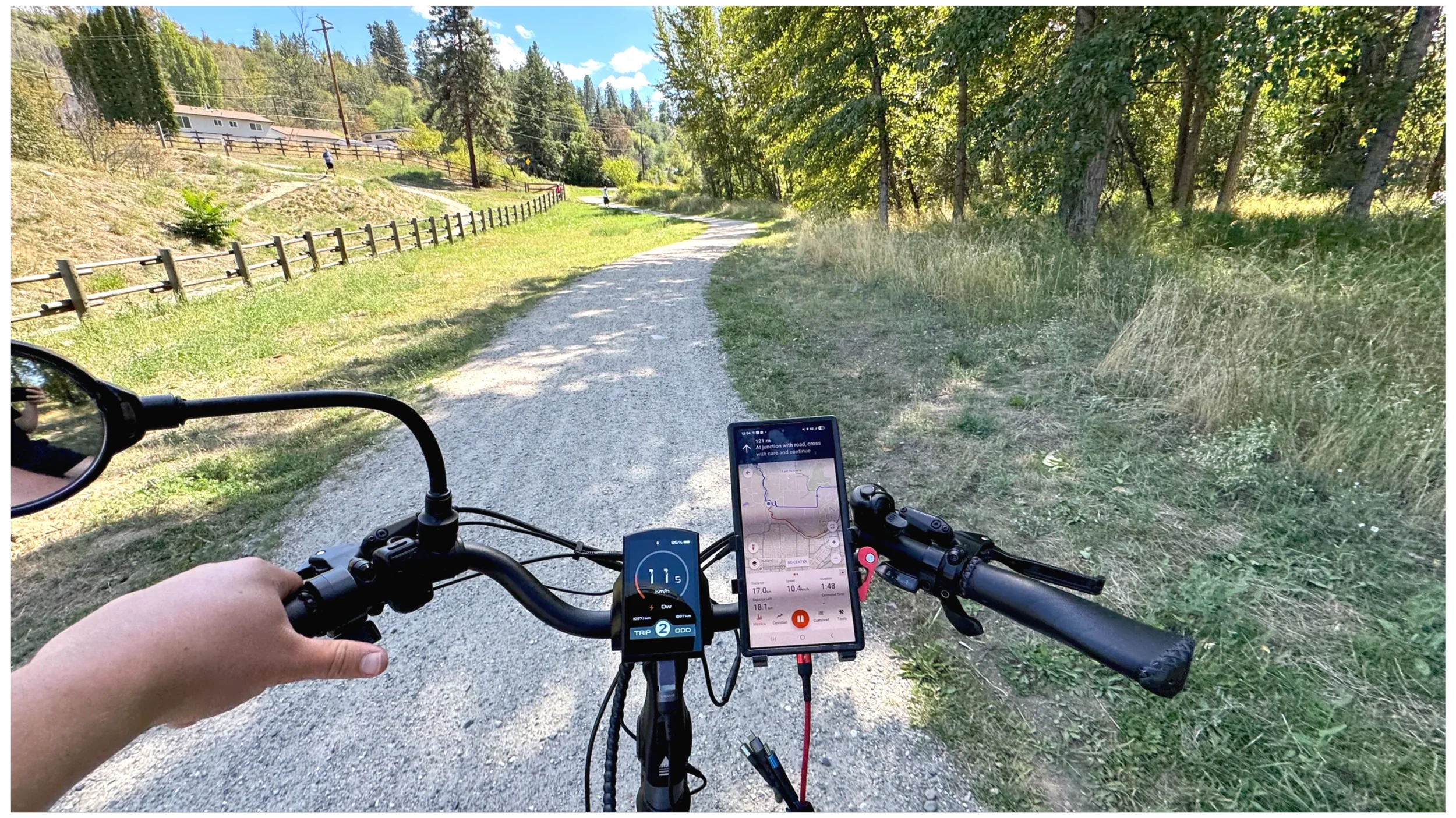 View from a bicycle dashboard showing a gravel trail winding through a grassy area with trees on the right and houses in the distance on the left, with a person riding a bicycle on the trail.
