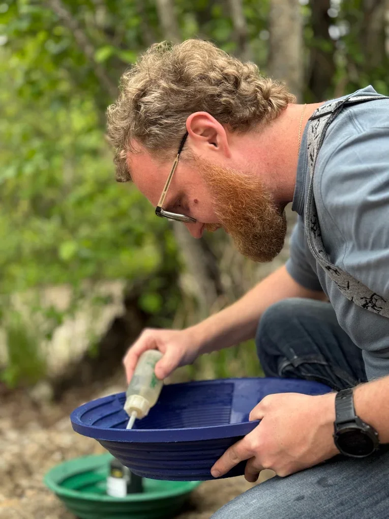 Man with glasses and a beard filling a blue bird feeder with nectar outdoors, surrounded by green trees.