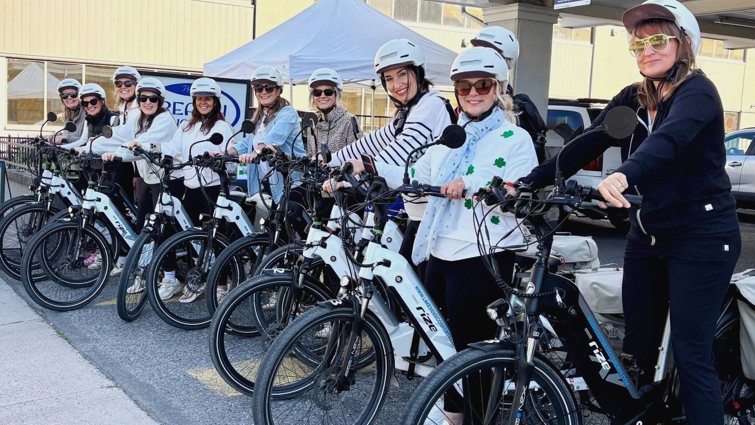 Group of women on electric Rize e-bikes wear Xnito helmets and sunglasses, standing in a line outdoors, ready for a ride in. Kelowna.