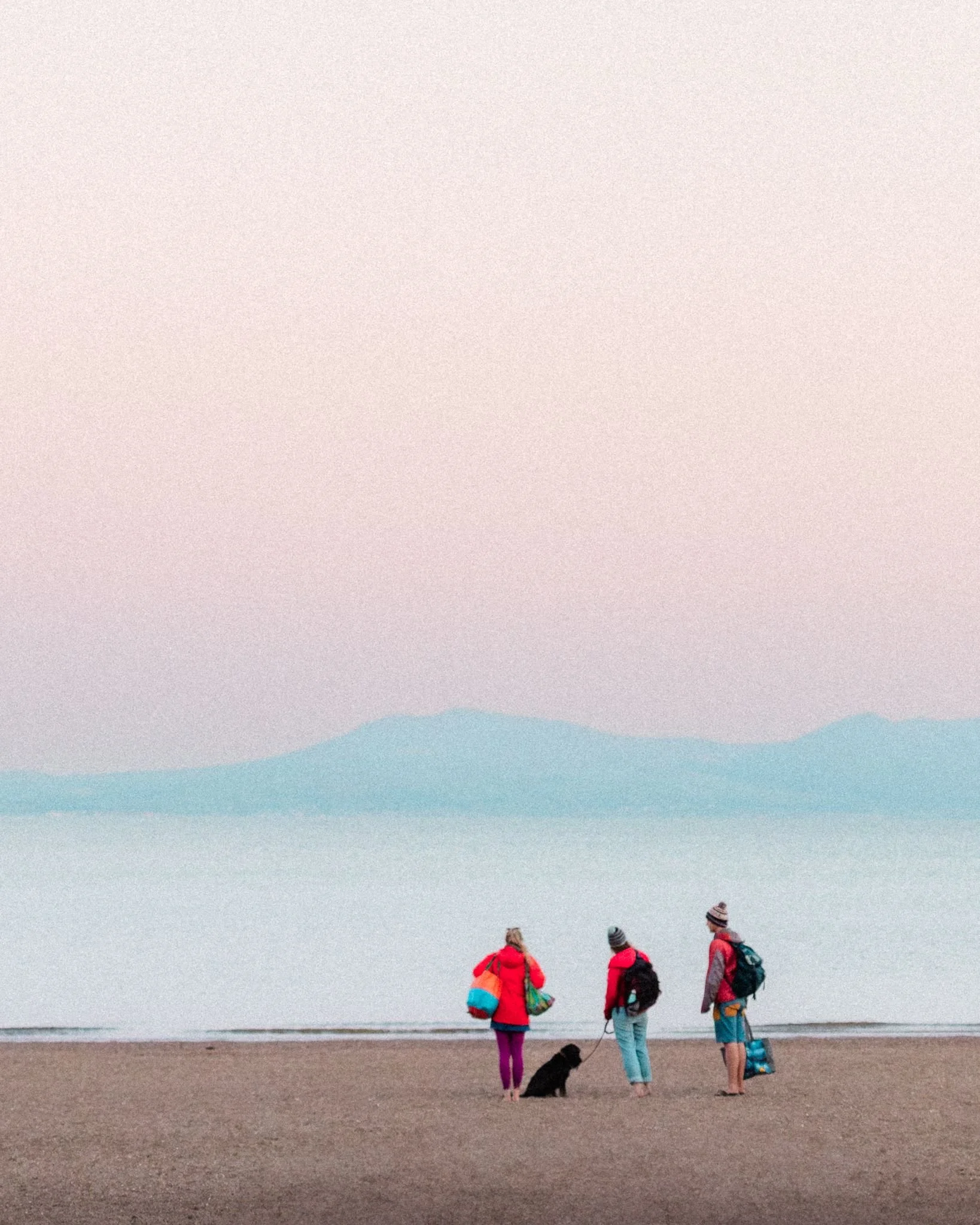 three figures stand on a sandy beach looking out at a dusky sunset over blue mountains in the distance.