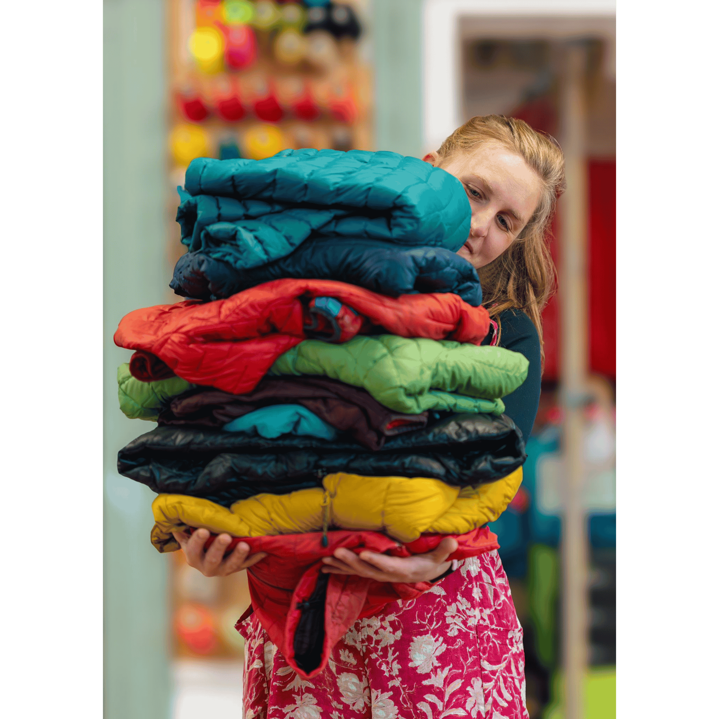 A smiling woman carries a large stack of down jackets in front of a thread board. The jackets have patches on them.