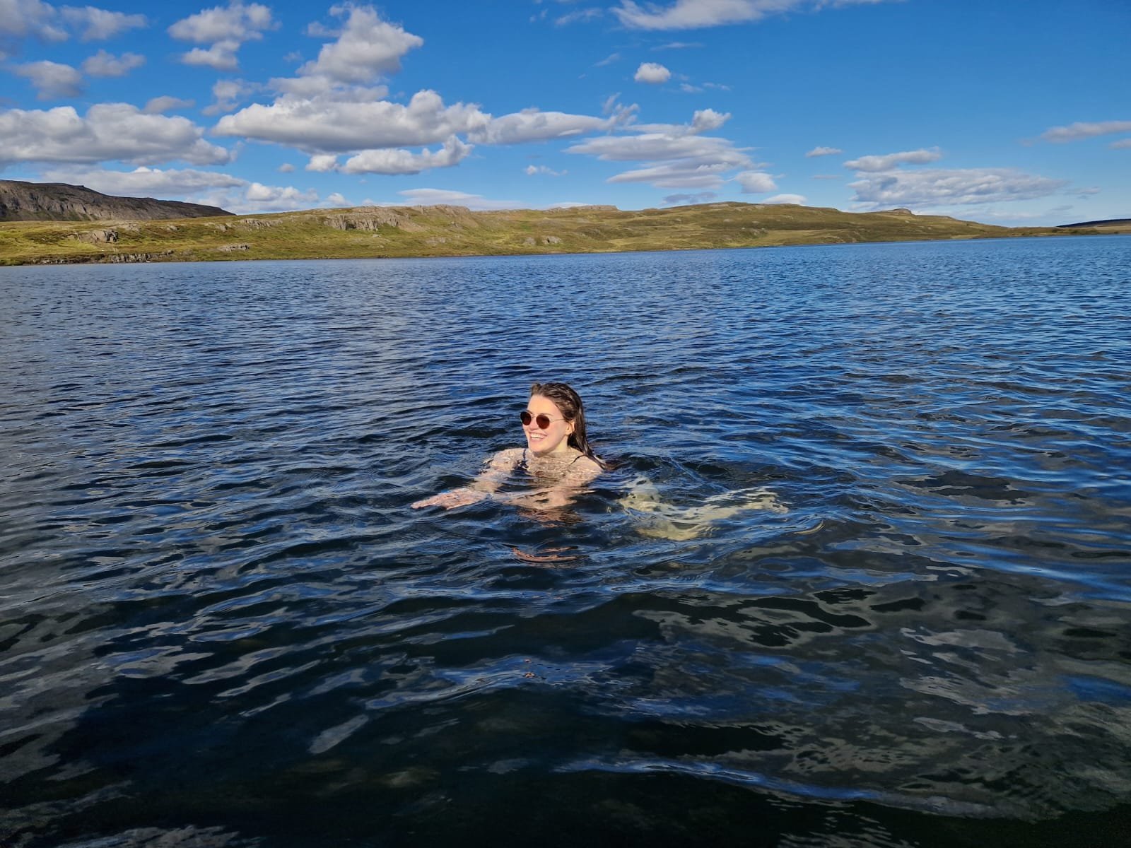 sunshine hits a woman wearing sunglasses who is wild swimming in a deep blue lake. The sky is clear with a few cumulous clouds, and green land can be seen behind her.