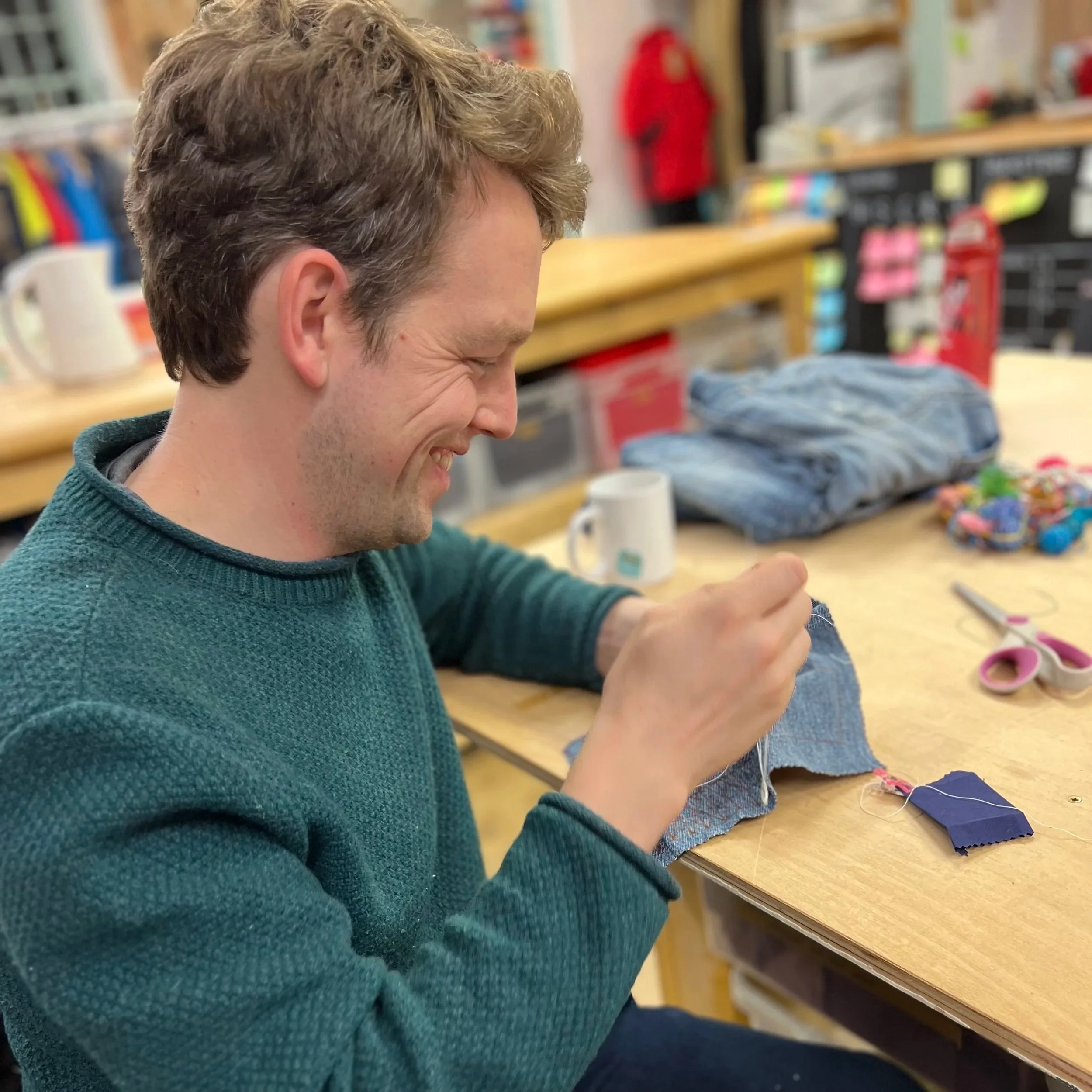 A man smiling and sewing fabric on a work table in a craft room, surrounded by scissors, fabric swatches, and other sewing materials.