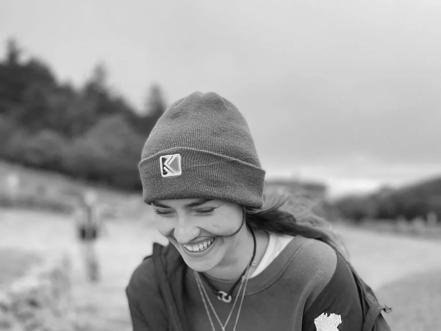 black and white image of a woman leaning forward and laughing, with a field and forest behind her. She is wearing a beanie and silver jewellery.