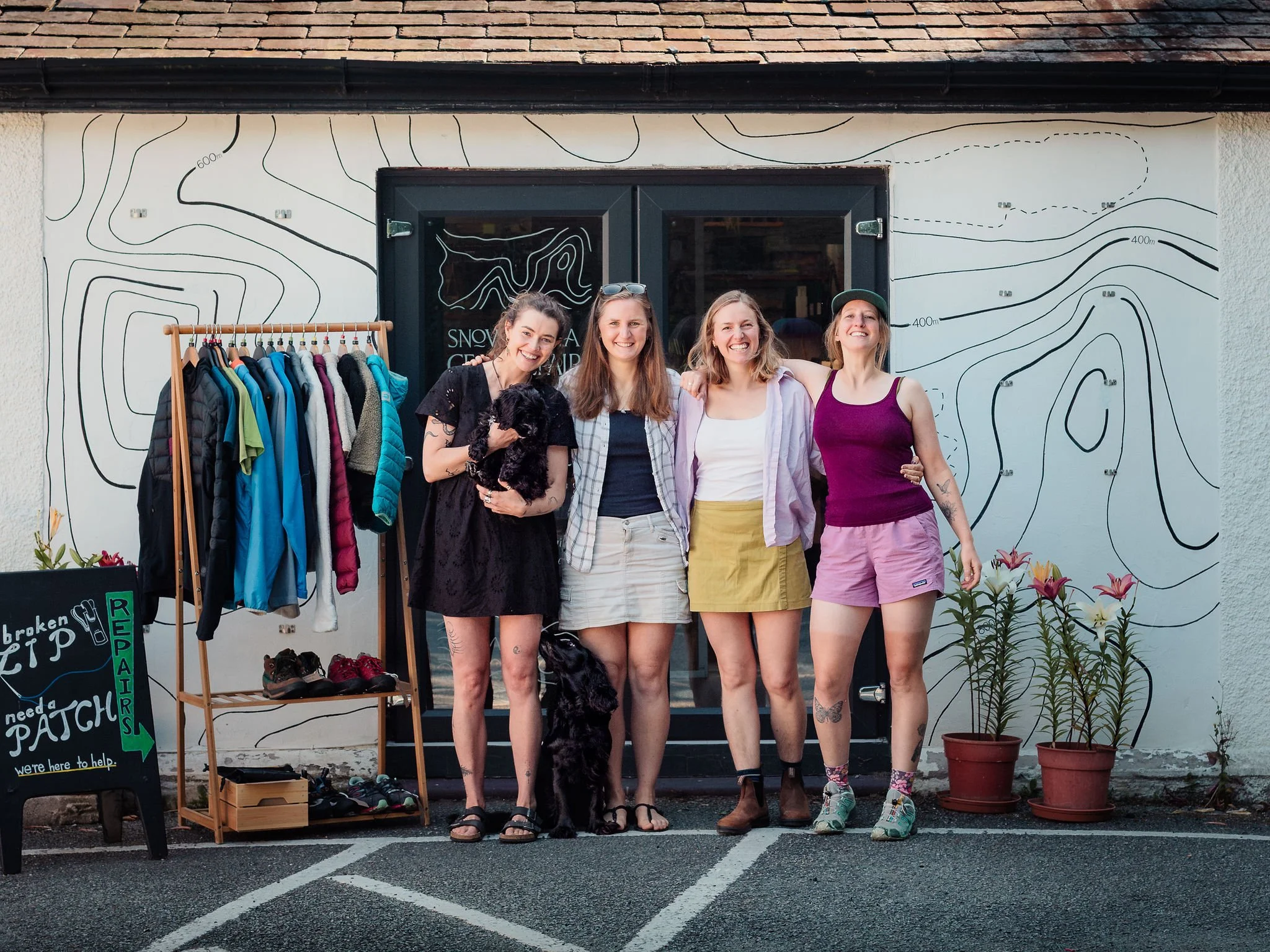 for women standing outside Snowdonia Gear Repair with two dogs and a sign that says 'broken zip, need a patch, we're here to help.' the building has contour lines painted on the outside.