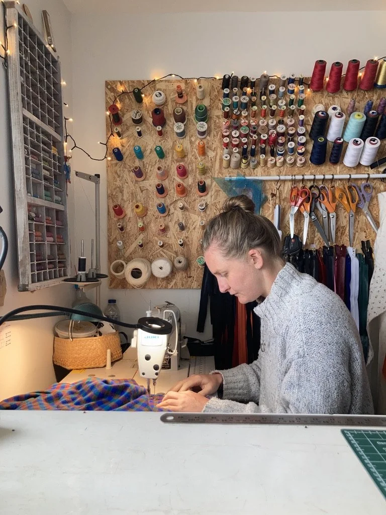 a woman stitching on an industrial machine at a home workshop, with a colourful threadboard in the background and selection of zippers hanging on the wall