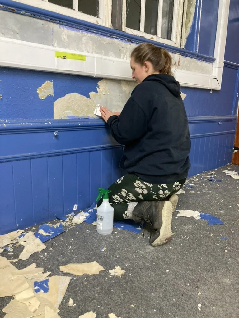 a woman kneels next to a wall from which she is scraping paint
