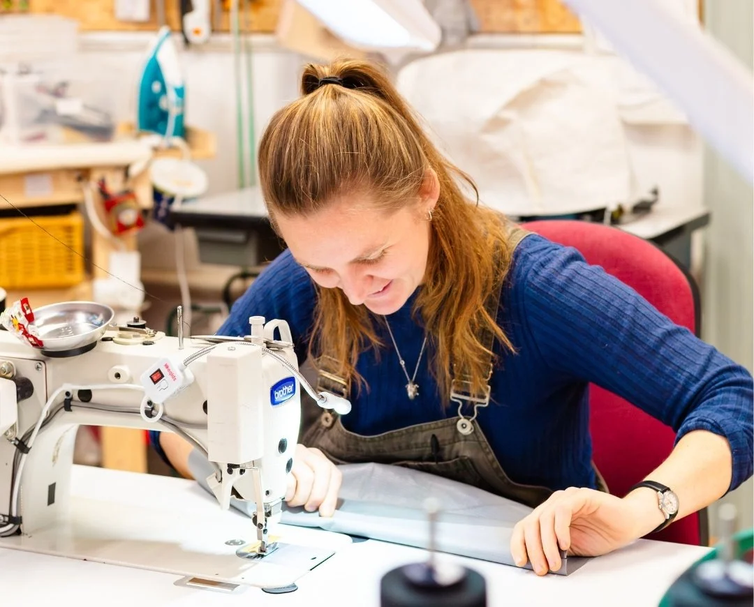 A woman in a purple turtleneck and dungarees prepares to stitch a roll-top back out of an upcycled camper awning. She's sitting at an industrial sewing machine in a workshop space.