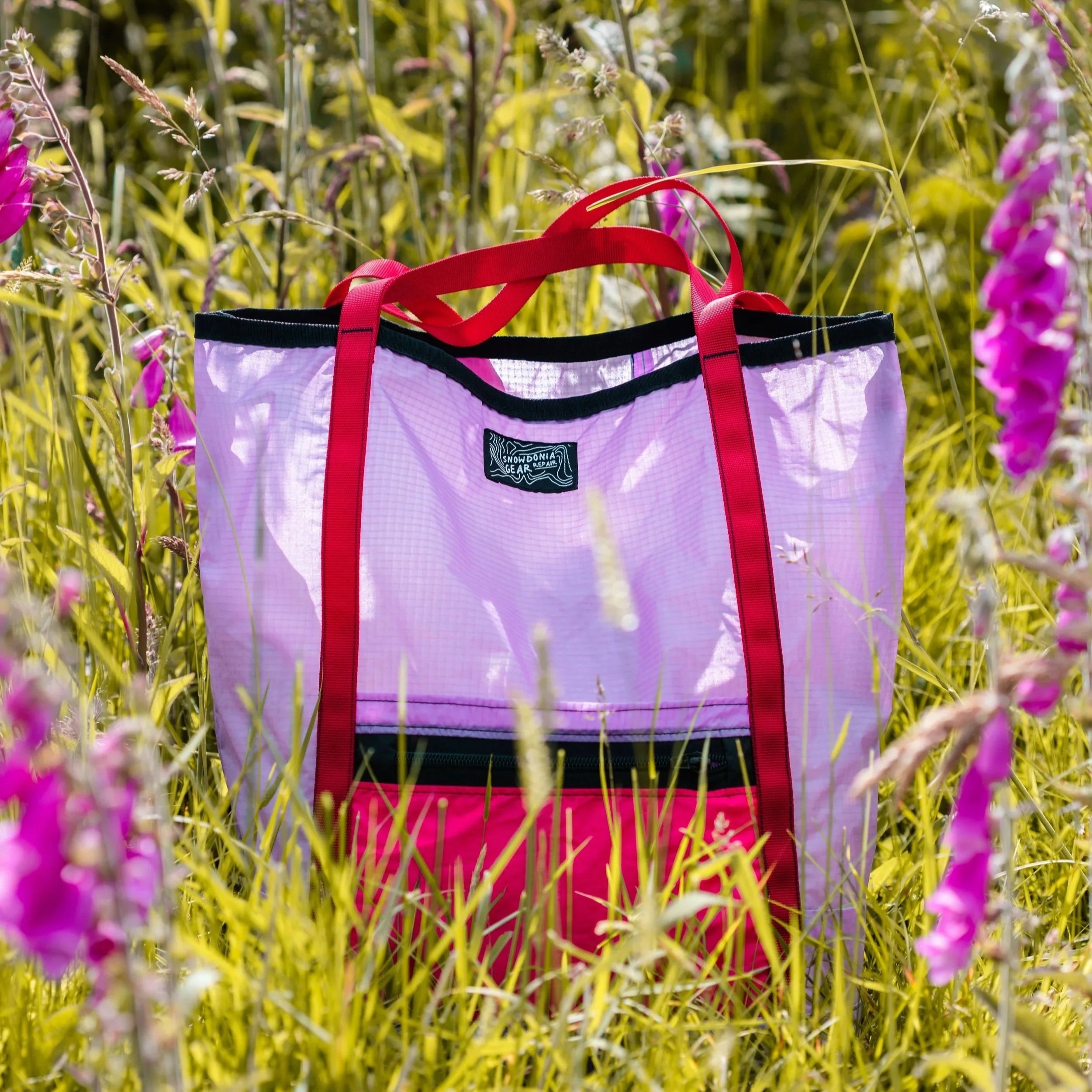 A pink and black tote bag with red straps sitting in a field of tall grass and purple flowers.