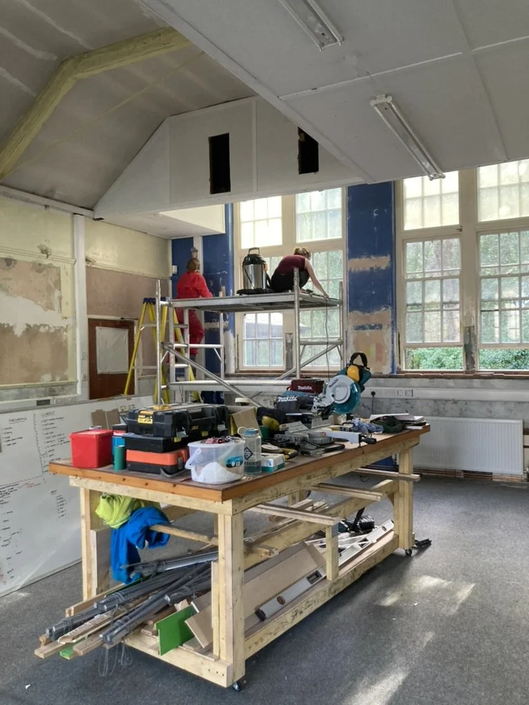 A large partially renovated room with a table in the middle covered in tools. Two figures stand on scaffolding in the background.