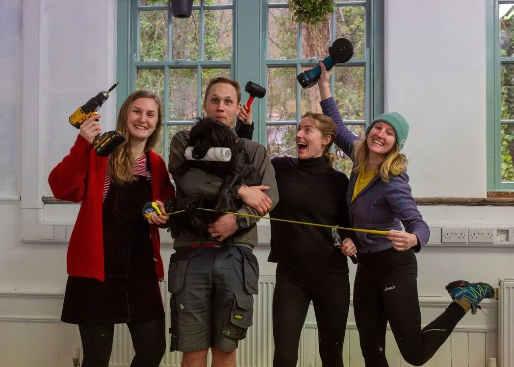 four people holding hand tools, like a drill, measuring tape, mallet, and paint roller making funny faces to celebrate the work they've done in a sparse looking room with forest outside the windows