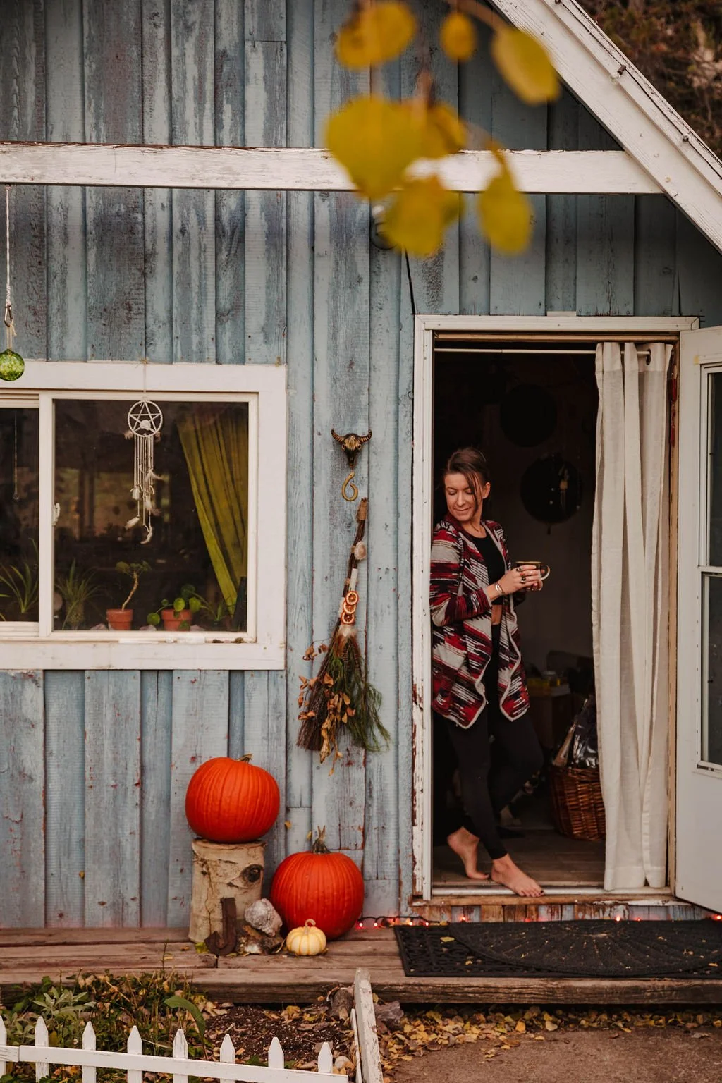 A woman standing barefoot at the doorway of a rustic wooden house, holding a mug, with autumn pumpkins and a small white pumpkin outside. Decor decorations include hanging herbs, a dream catcher, and a bull skull with a ring.