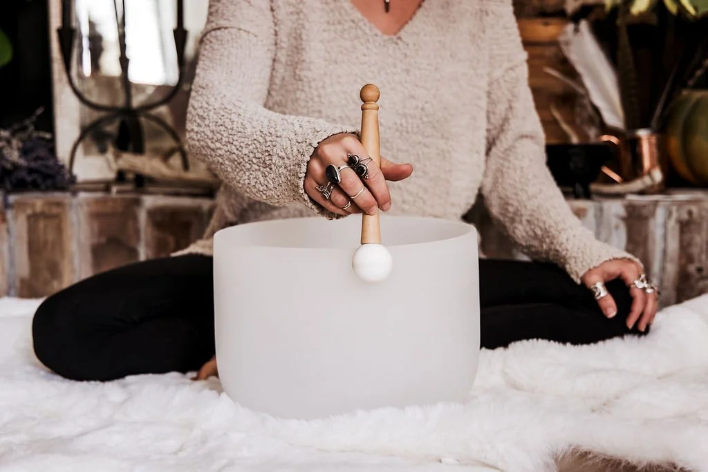 Person sitting cross-legged on a cozy white blanket, playing a white singing bowl with a wooden mallet, wearing rings and a beige sweater.