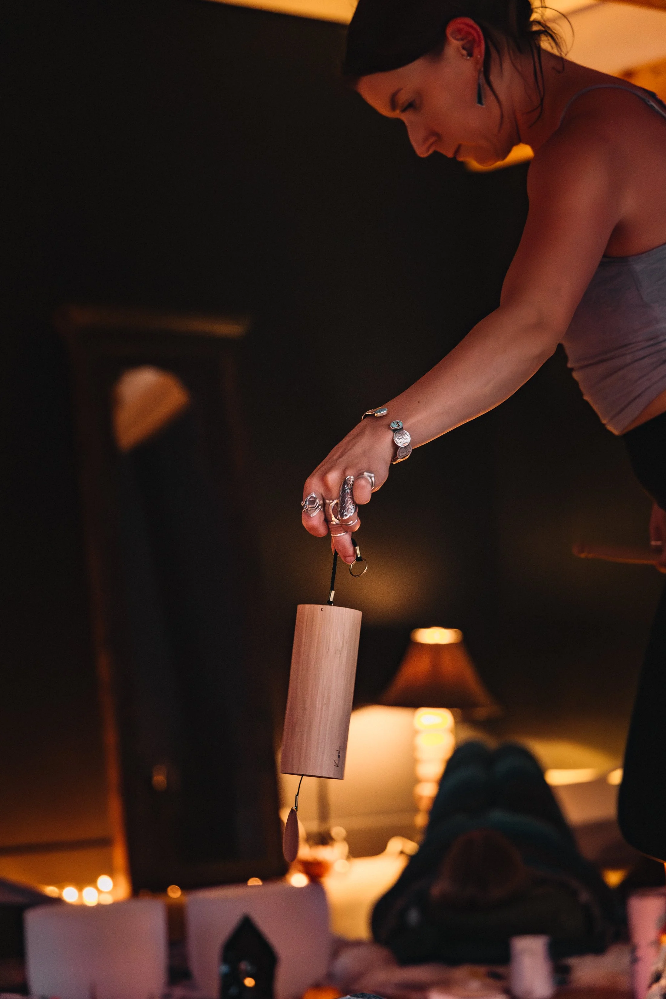A woman wearing jewelry and a tank top is hanging a wind chime from the ceiling in a dimly lit room with warm lighting.