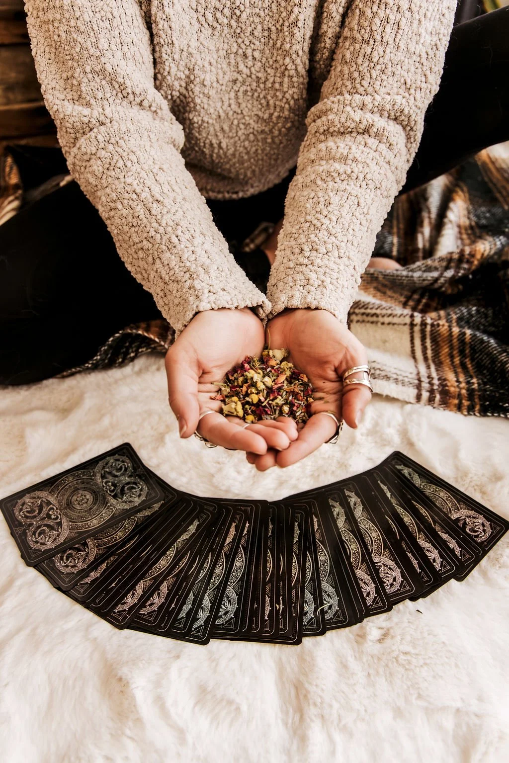 Person in a cozy beige sweater sitting on a blanket, holding a handful of dried herbs or flowers, with tarot cards spread out on a soft surface in front.