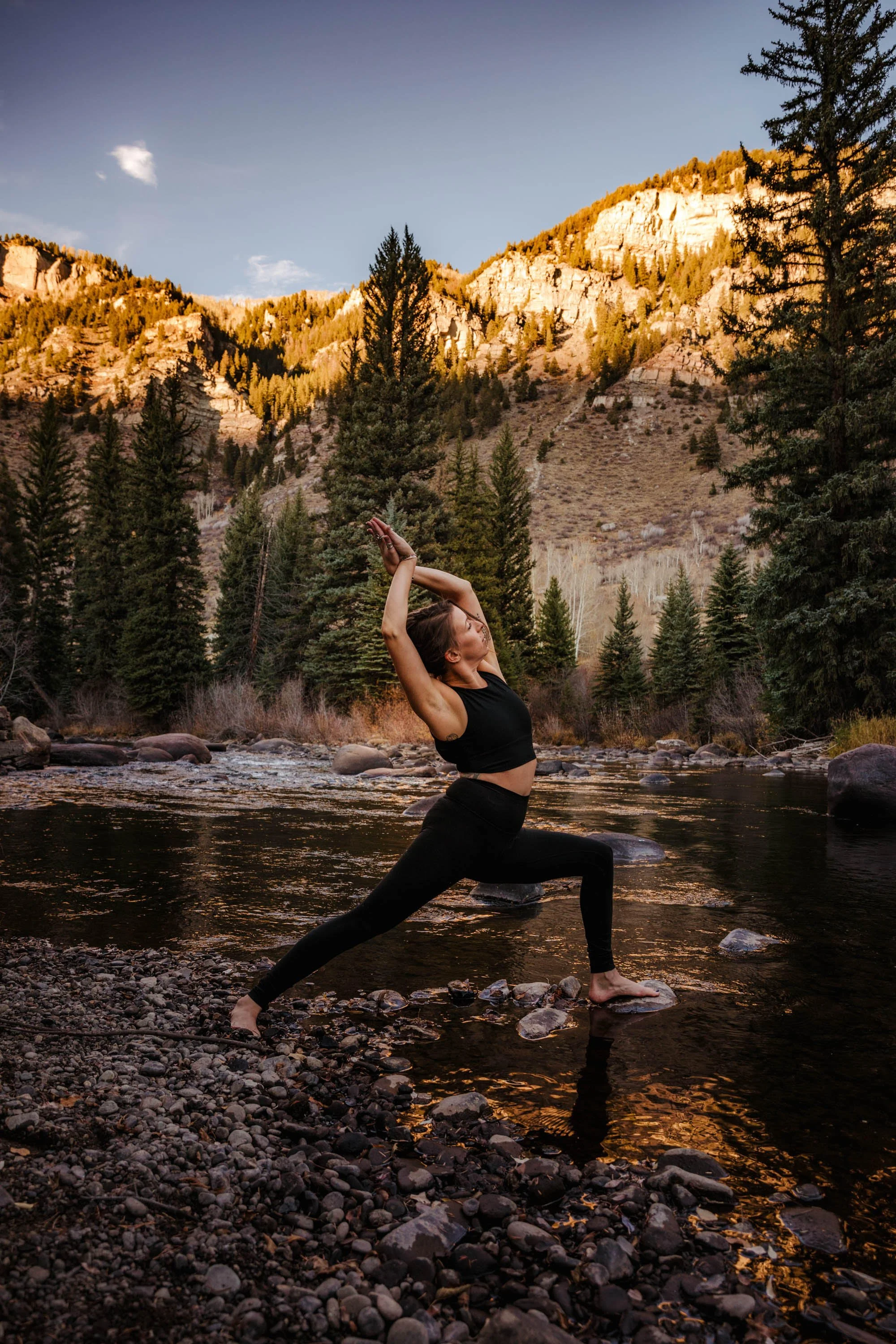 A woman practicing yoga in a lunge pose in a river with mountainous landscape and pine trees in the background during sunset.
