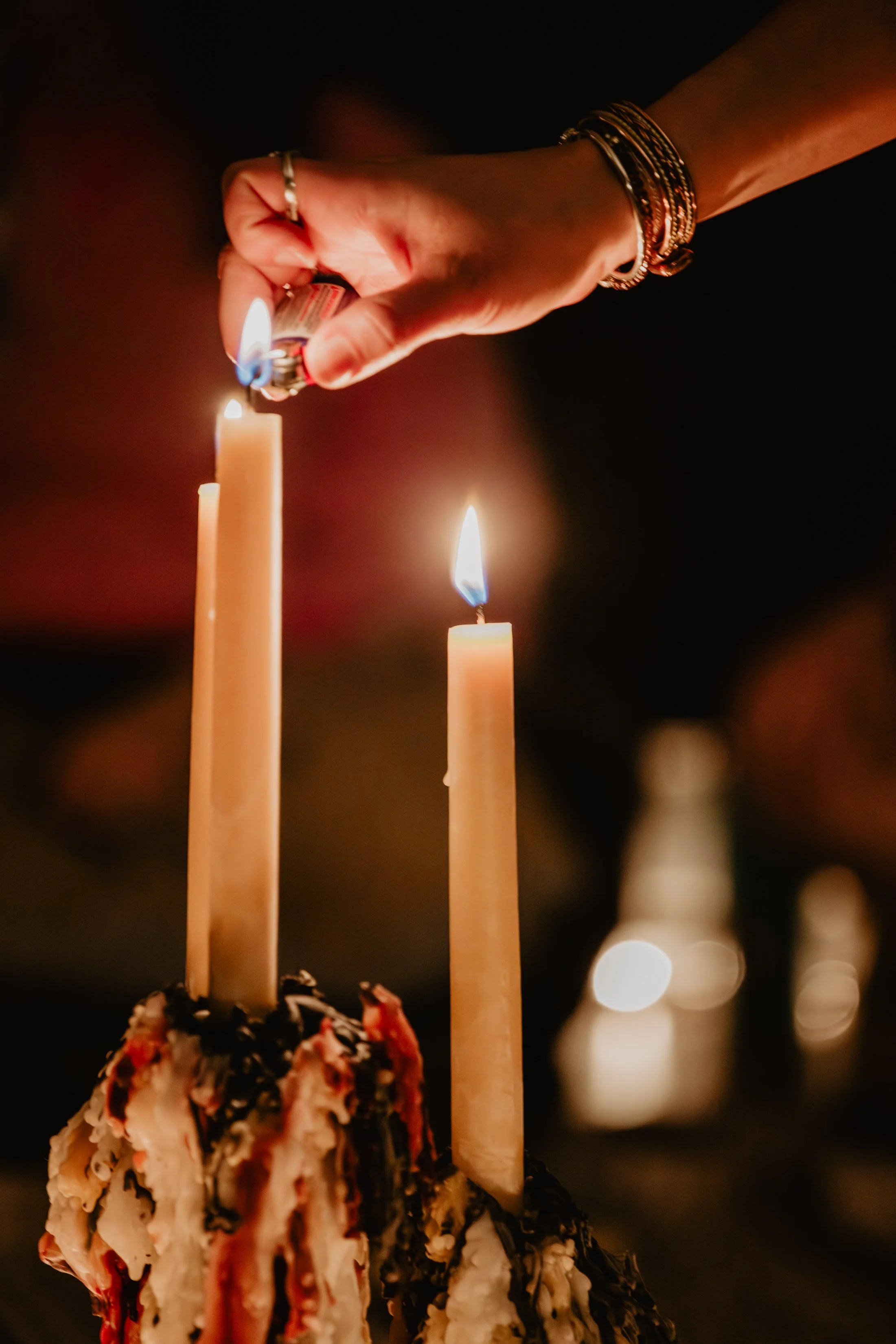 A hand holding a lighter igniting a candle wick. Two candles are lit with flames, and the hand is adorned with jewelry, including rings and bracelets. The background is dark and blurred.