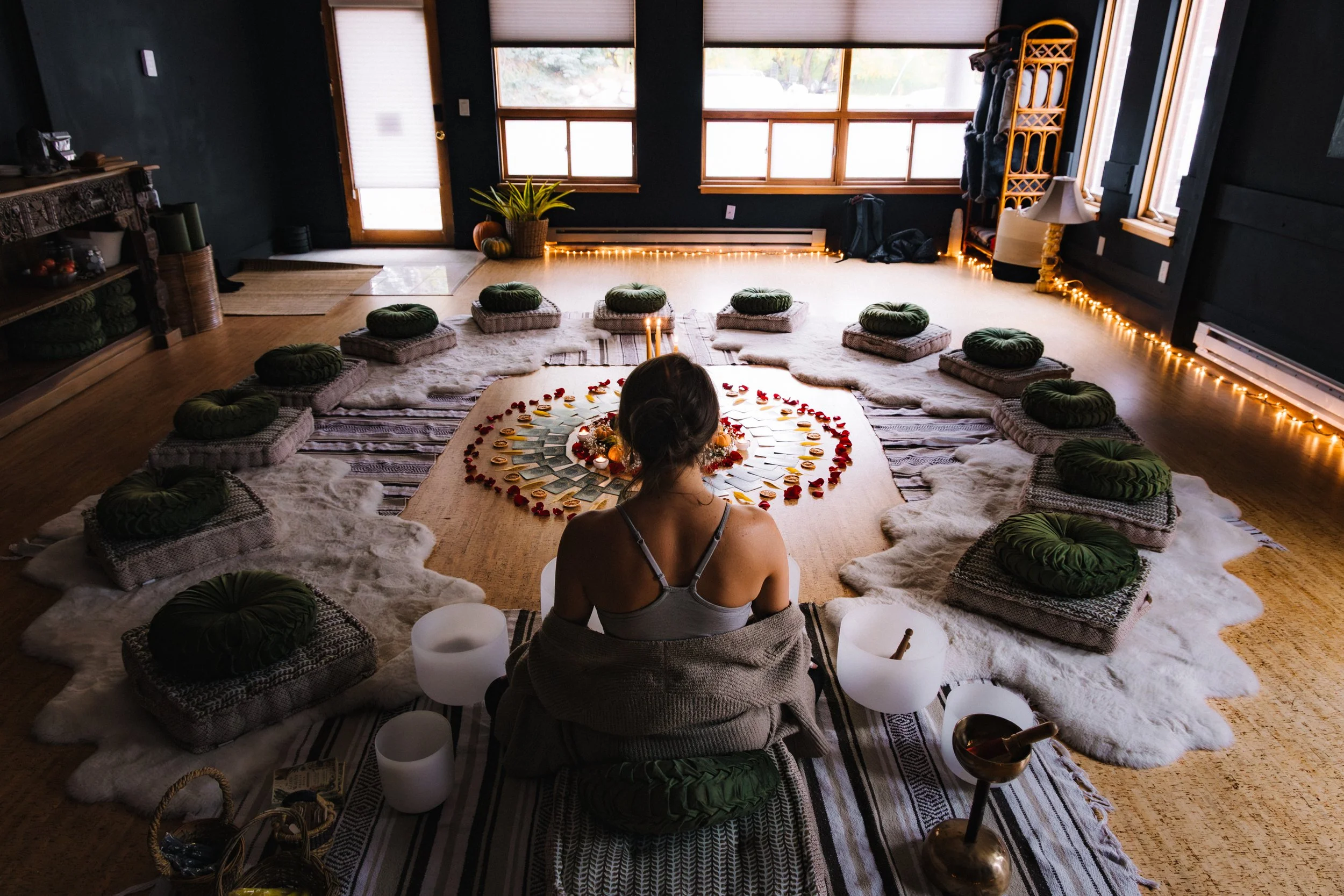 A cozy indoor space decorated for a celebration with a large circular arrangement of cards or photos, surrounded by green pillows on the floor, room illuminated by string lights, and a person sitting cross-legged facing away.