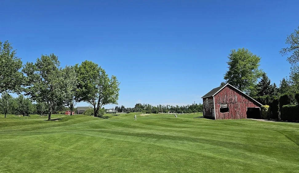 A golf course with green grass, trees, a red barn, and a clear blue sky.