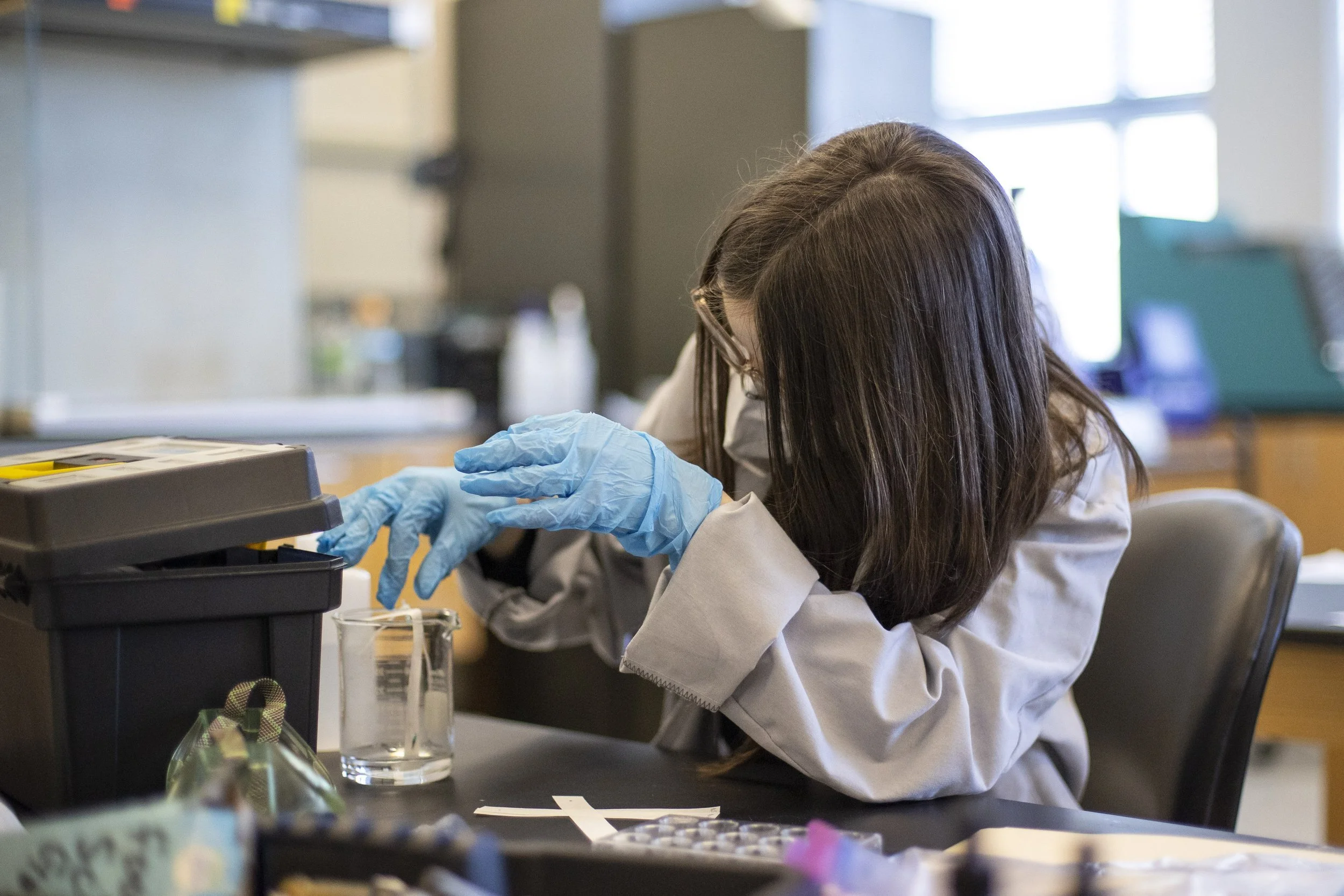 Person in a lab coat and gloves working in a laboratory with a beaker and equipment on the table
