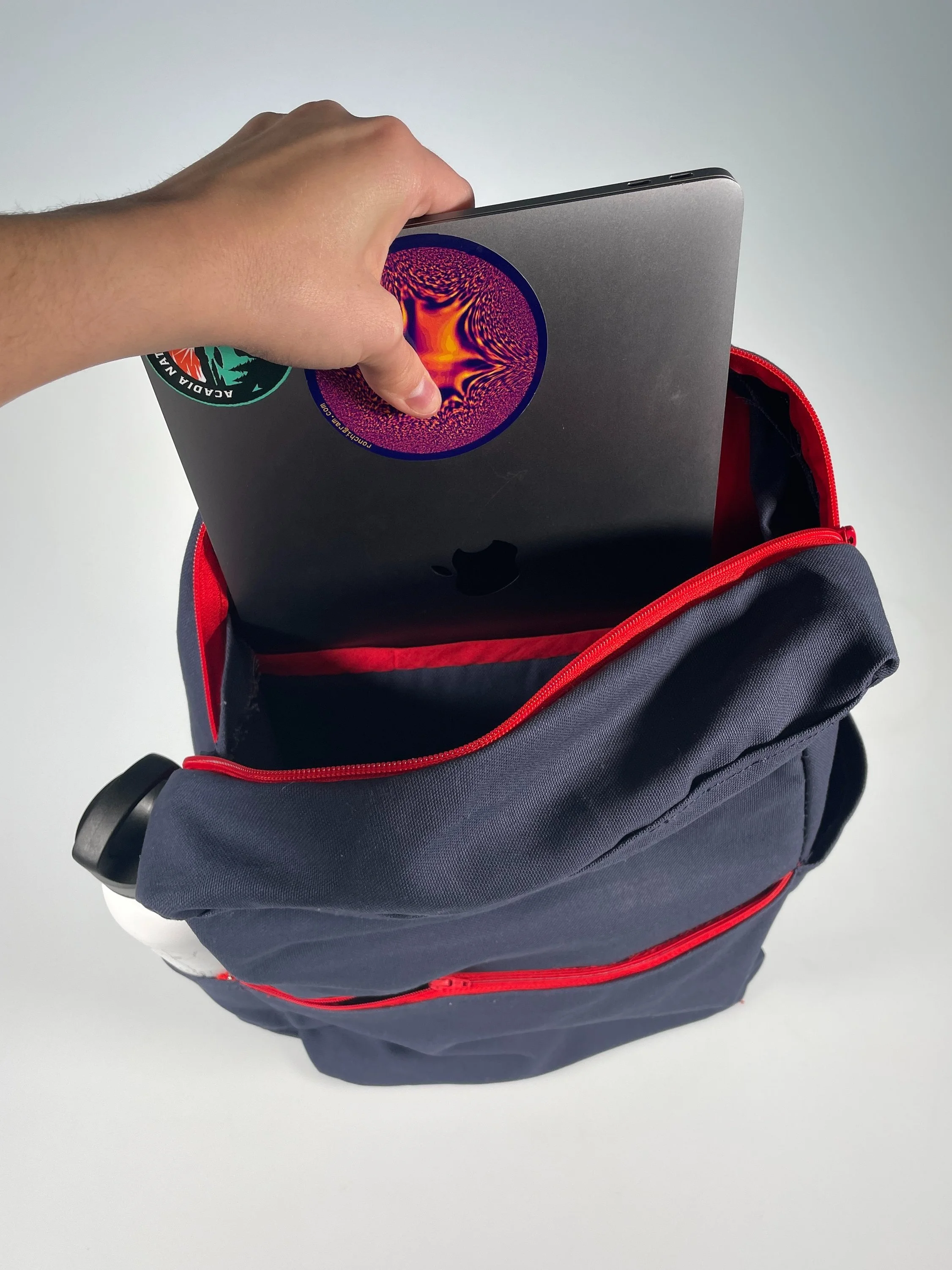 Person putting a laptop with stickers into a blue backpack with red zippers and a water bottle side pocket.