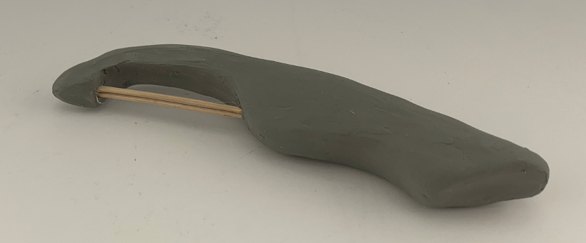 Handcrafted clay sculpture with an elongated shape featuring a wooden stick embedded inside, lying on a neutral background.
