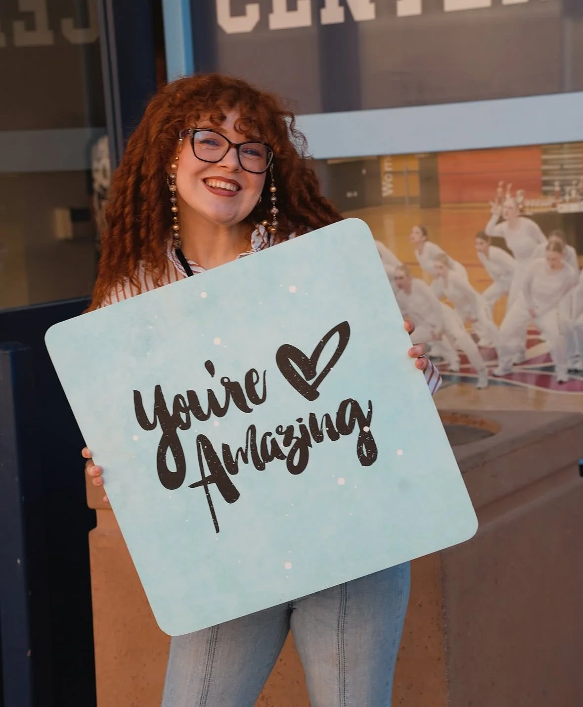 A smiling woman with curly red hair and glasses holding a sign that says 'You're Amazing' with a heart. She is standing outside a building with a large window and a motivational poster of a group of women in white sportswear in the background.