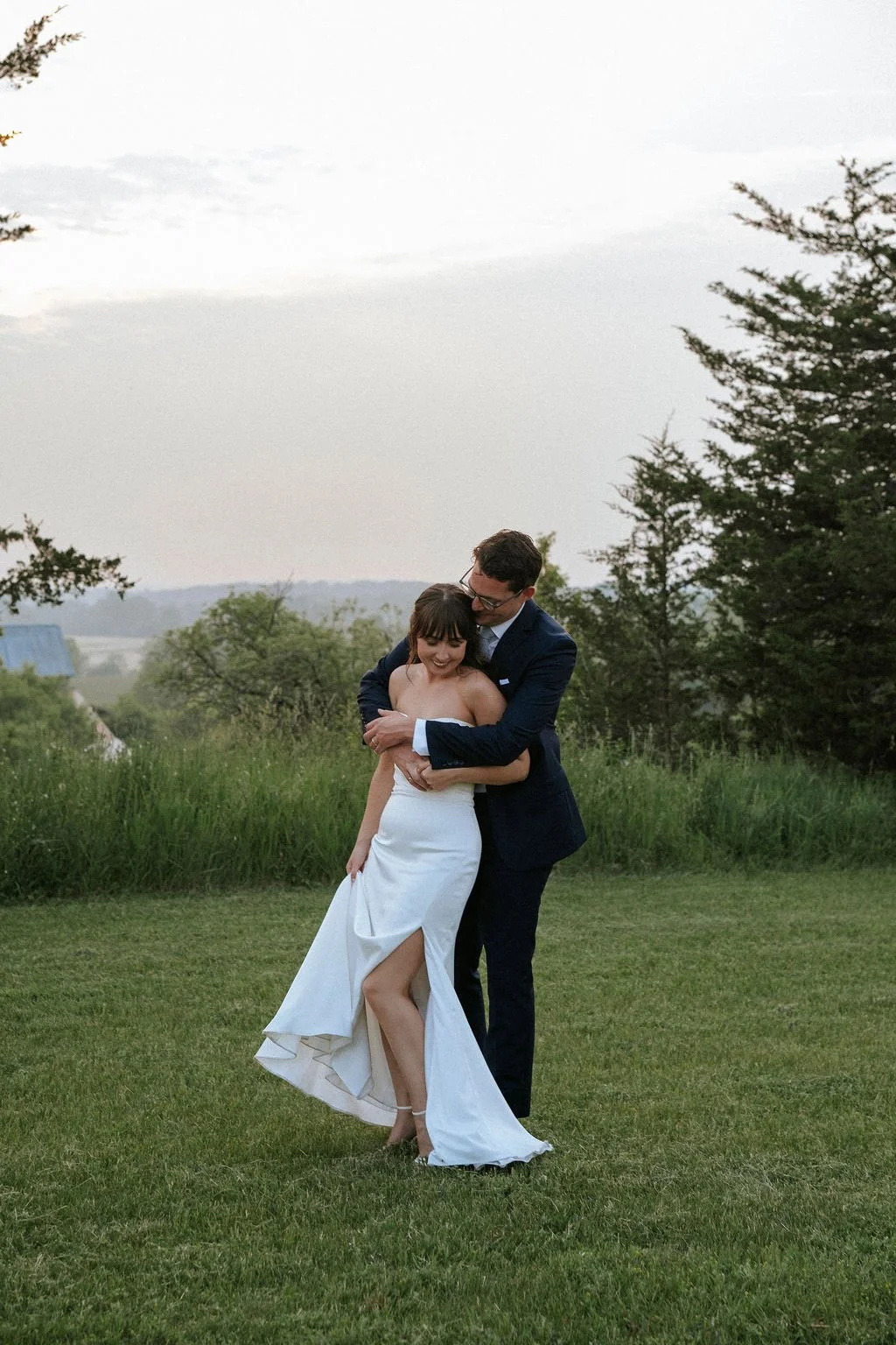 A newlywed couple embracing outdoors on a grassy field with trees and a cloudy sky in the background.