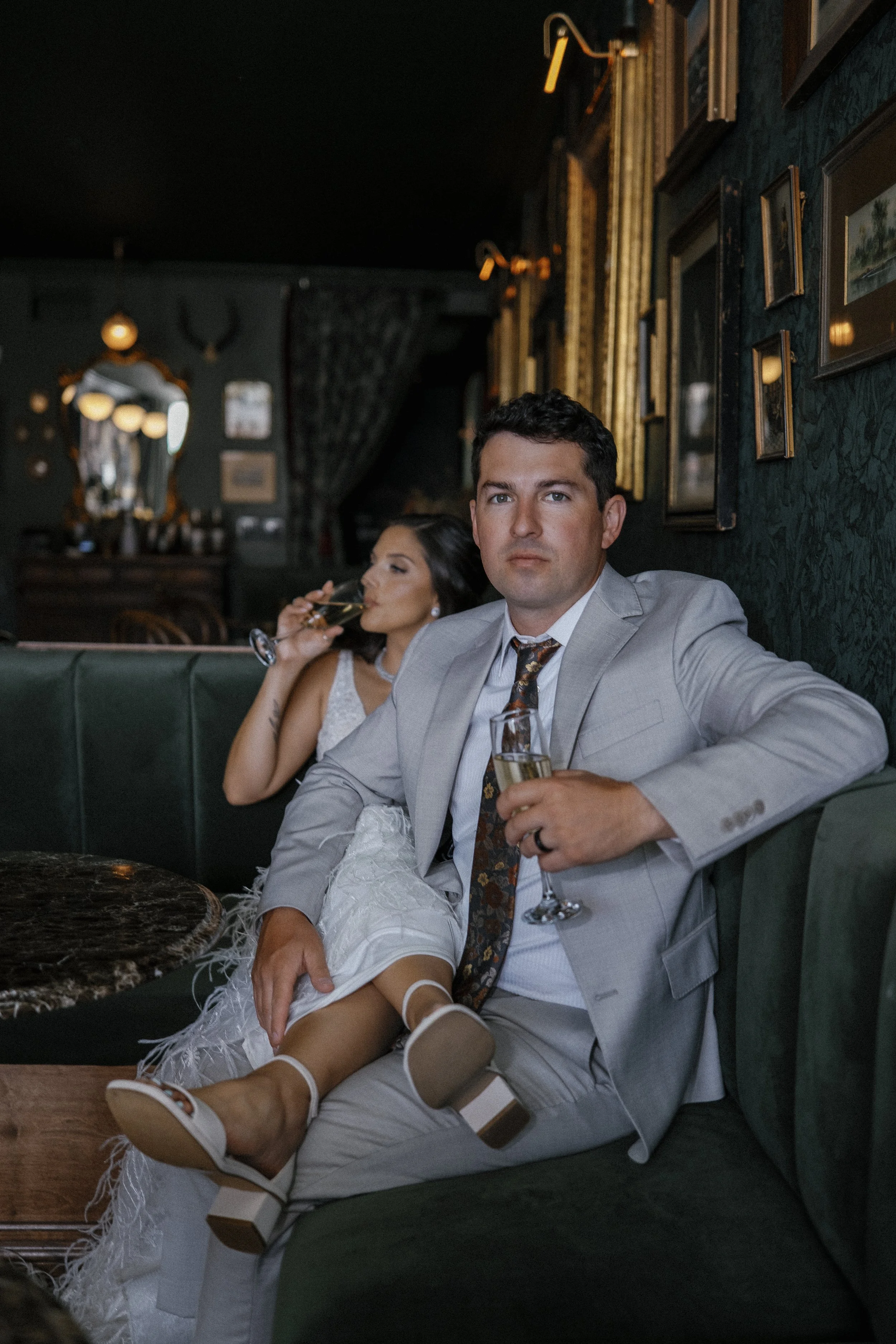 A man in a light gray suit and a woman in a white dress are sitting on a green leather booth in a dimly lit, vintage-style bar or restaurant. The man is looking at the camera holding a glass of champagne, while the woman is drinking from a similar gl