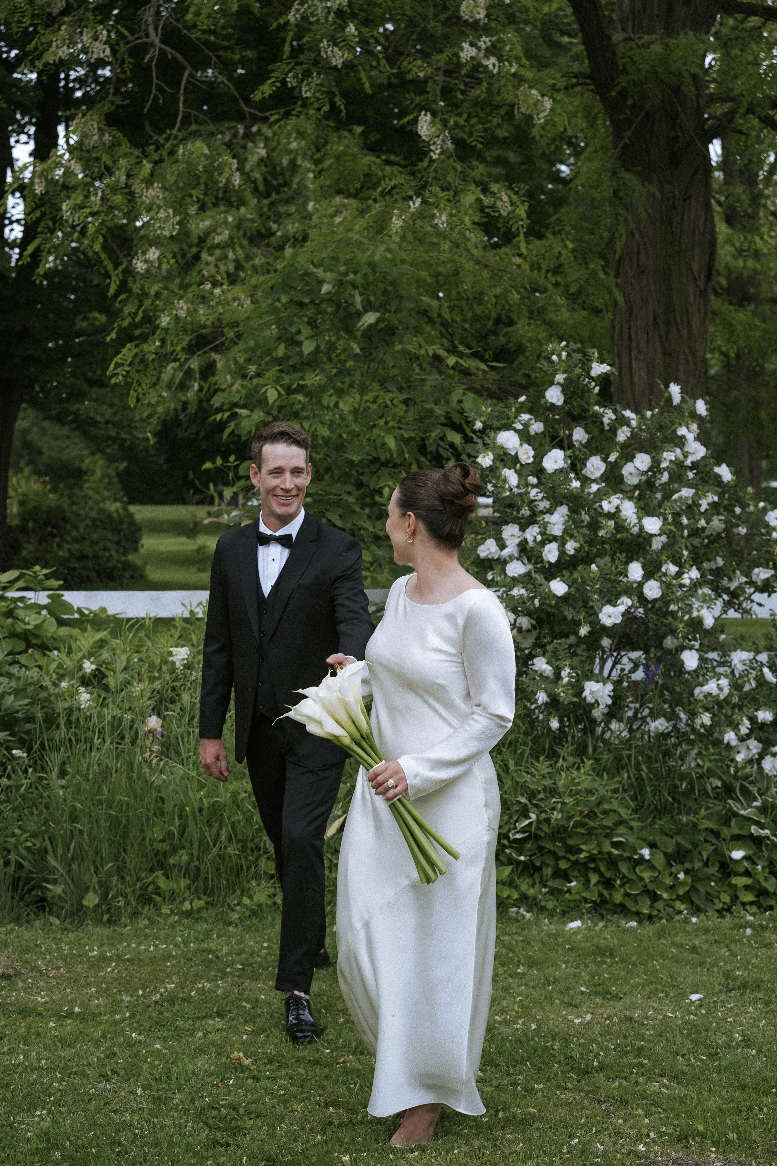 A bride and groom walking together outdoors, smiling. The bride is holding a bouquet of white calla lilies and is dressed in a long white gown. The groom is dressed in a black tuxedo with a bow tie. They are surrounded by green trees and white flowering bushes.