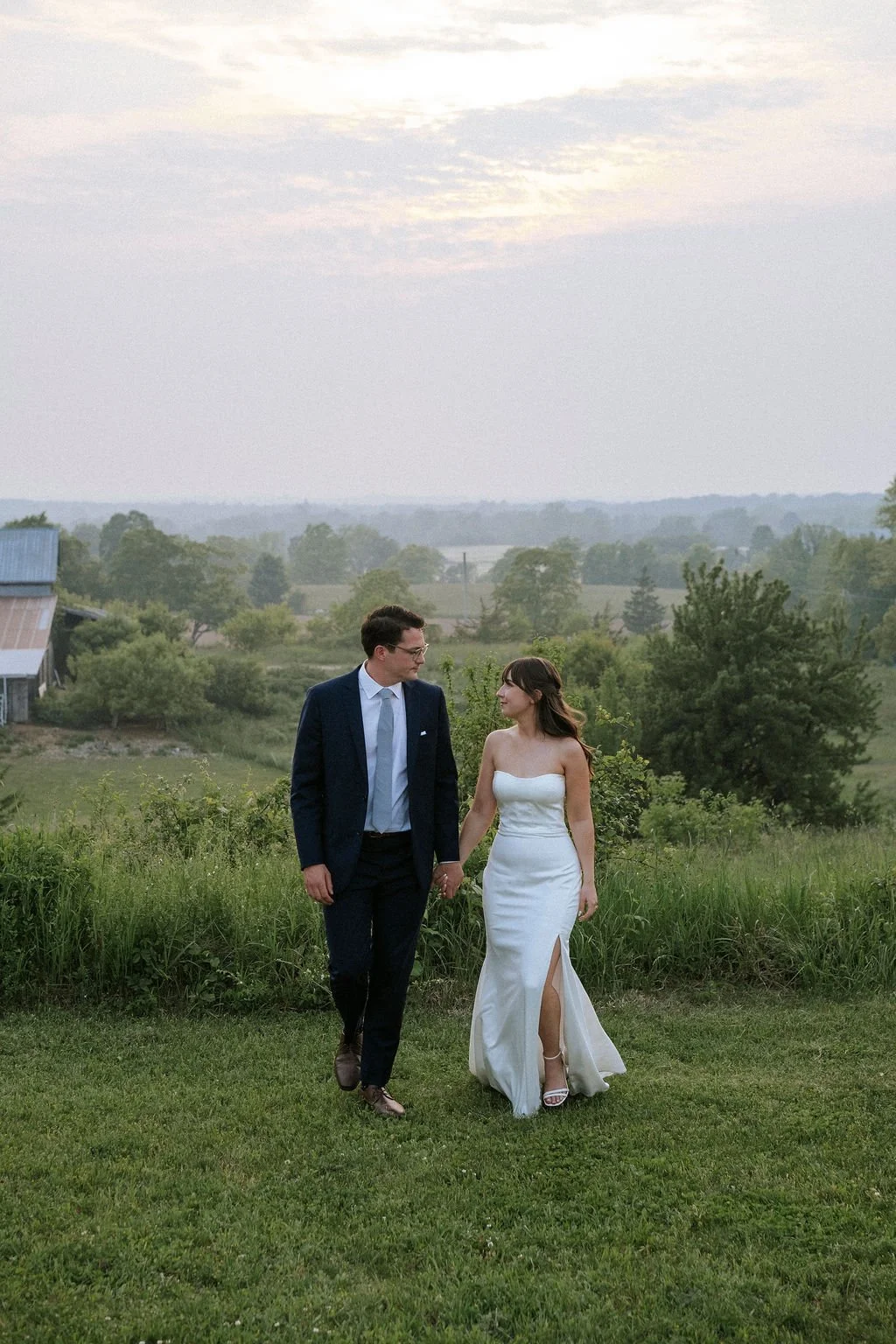 A couple in wedding attire walking hand in hand on a grassy field with trees and a cloudy sky in the background.