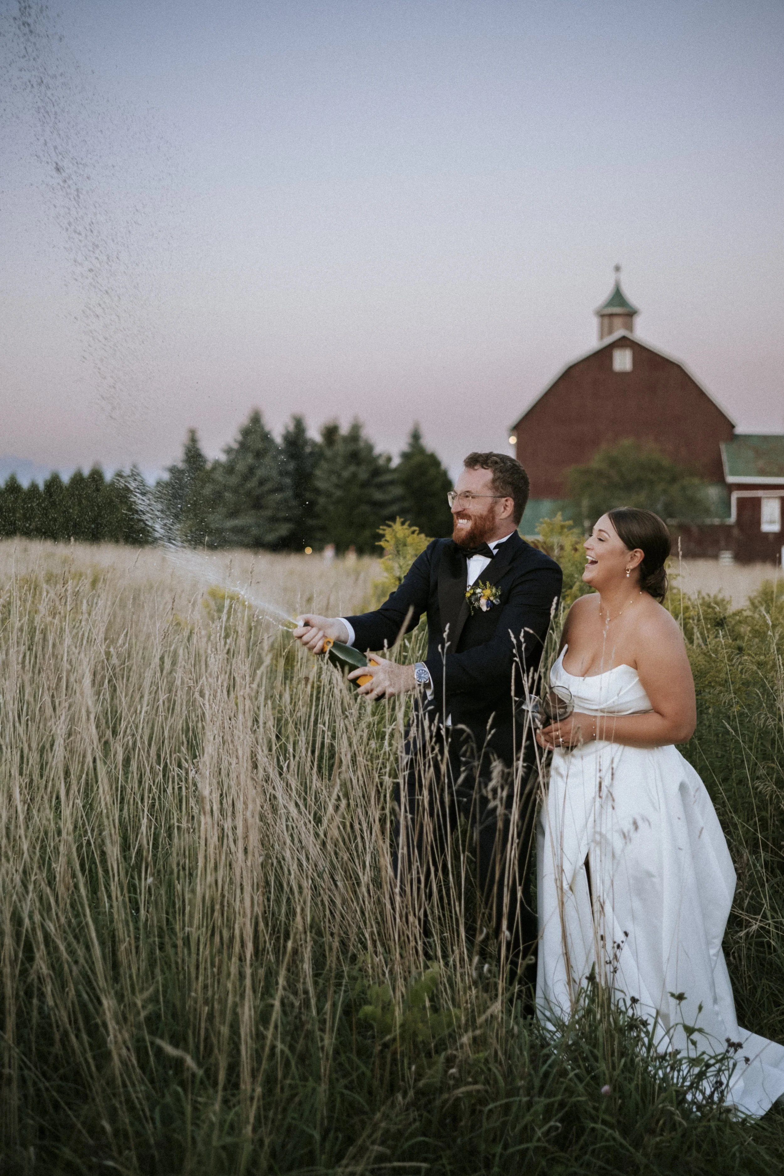 A newlywed couple celebrating outdoors in a field with tall grass, popping a bottle of champagne. The man has a beard and is wearing a tuxedo, while the woman is in a white wedding gown. A red barn and trees are visible in the background during dusk.