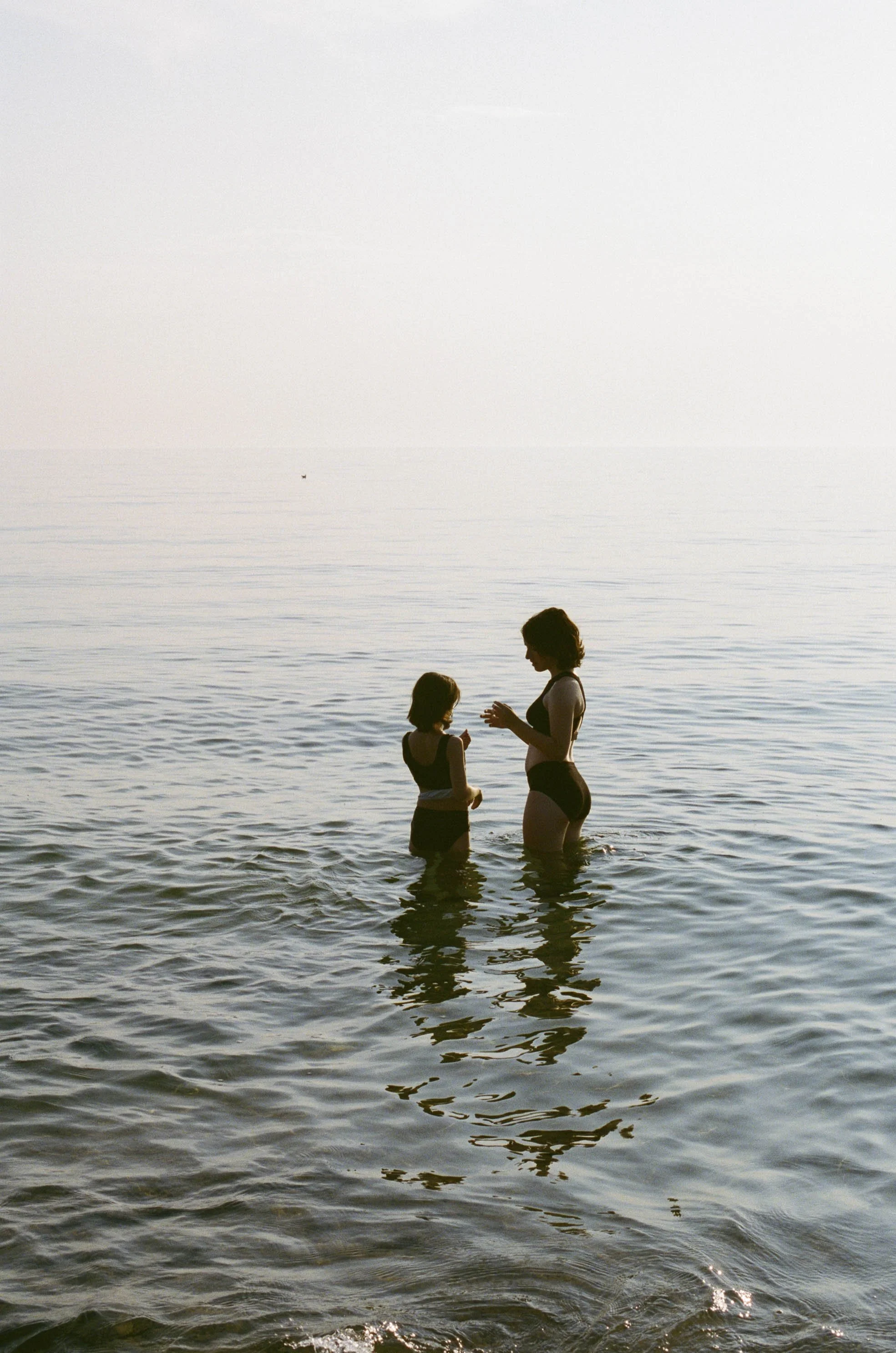 Two women in swimsuits standing in shallow water at the beach, having a conversation, with the ocean and sky in the background.