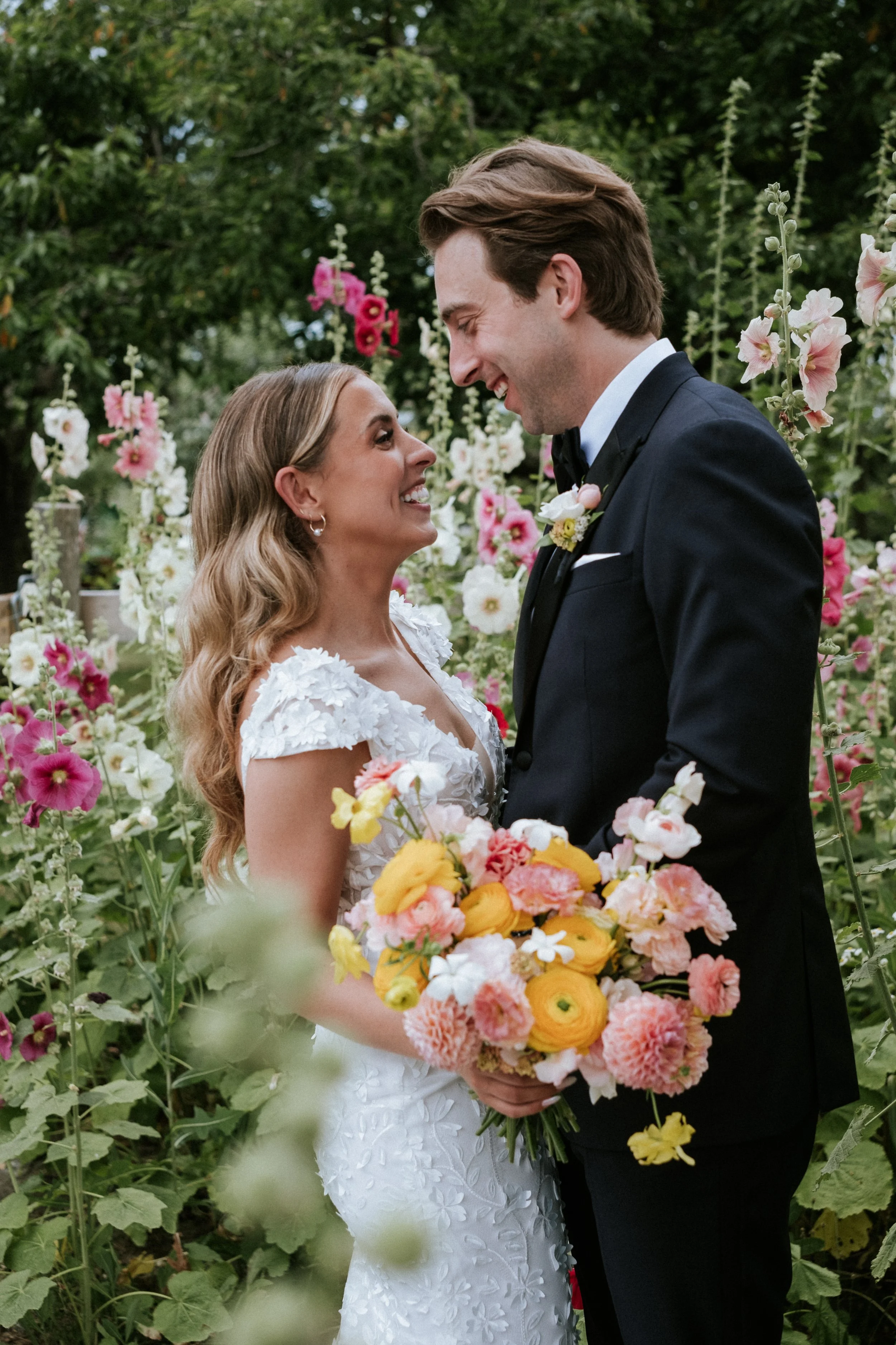 A bride and groom stand close together in a garden filled with pink, white, and yellow flowers, smiling at each other during their wedding ceremony.