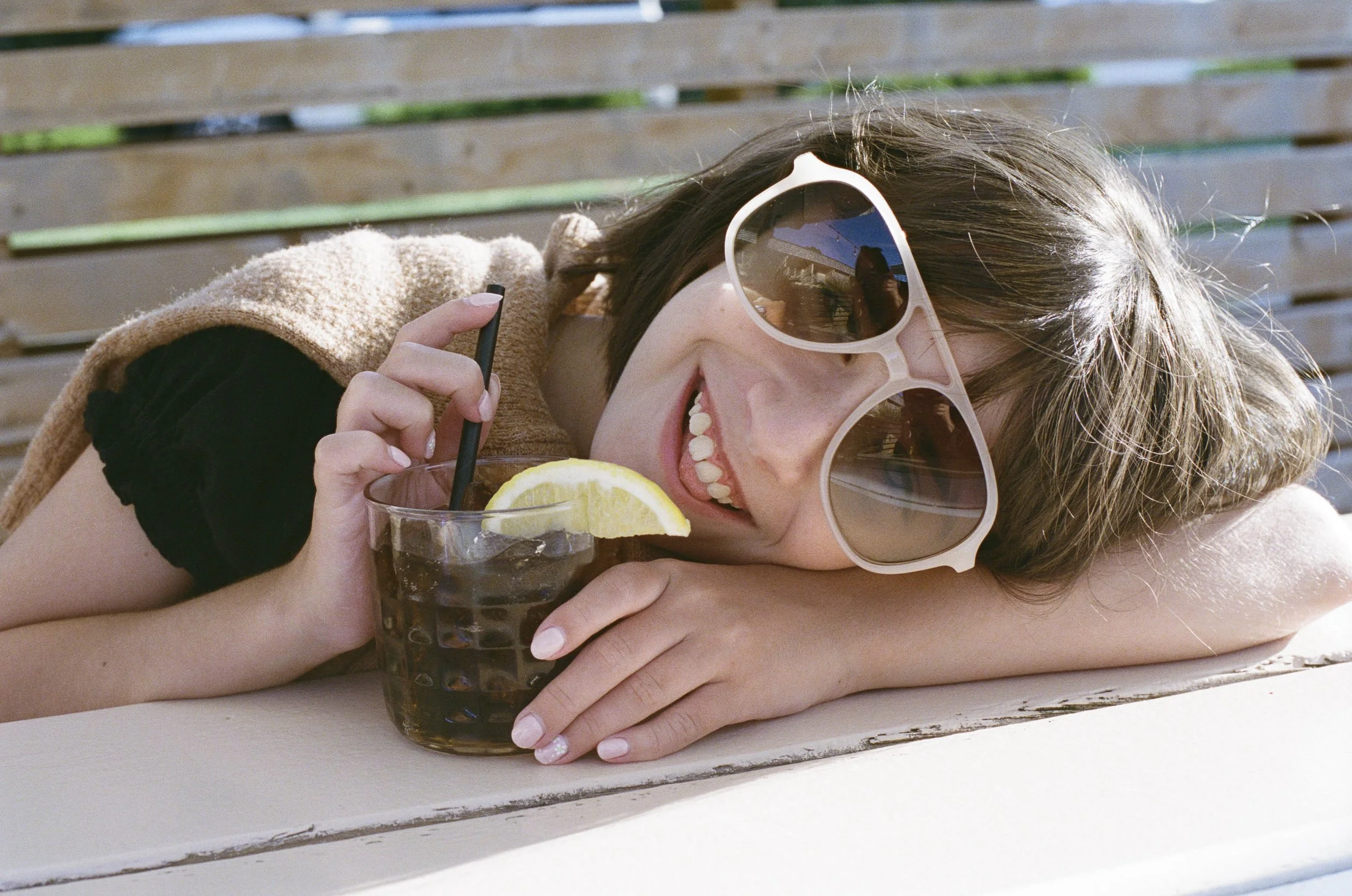 Young woman lounging outdoors wearing large sunglasses, resting her head on her arm, holding a glass of iced cola with a lemon wedge.