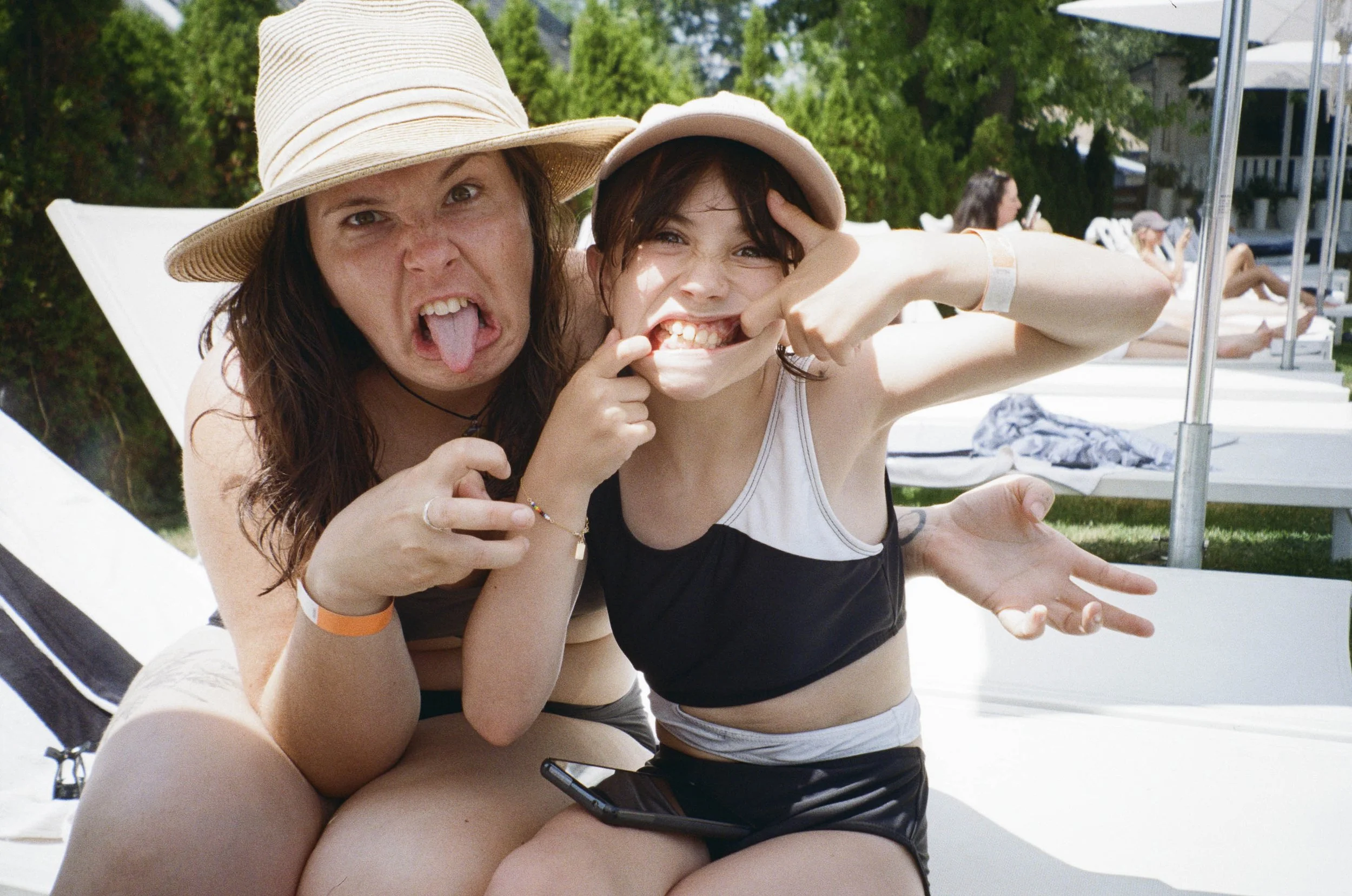 Two young women making funny faces at a poolside on a sunny day, both wearing swimsuits and hats, with lounge chairs and umbrellas in the background.