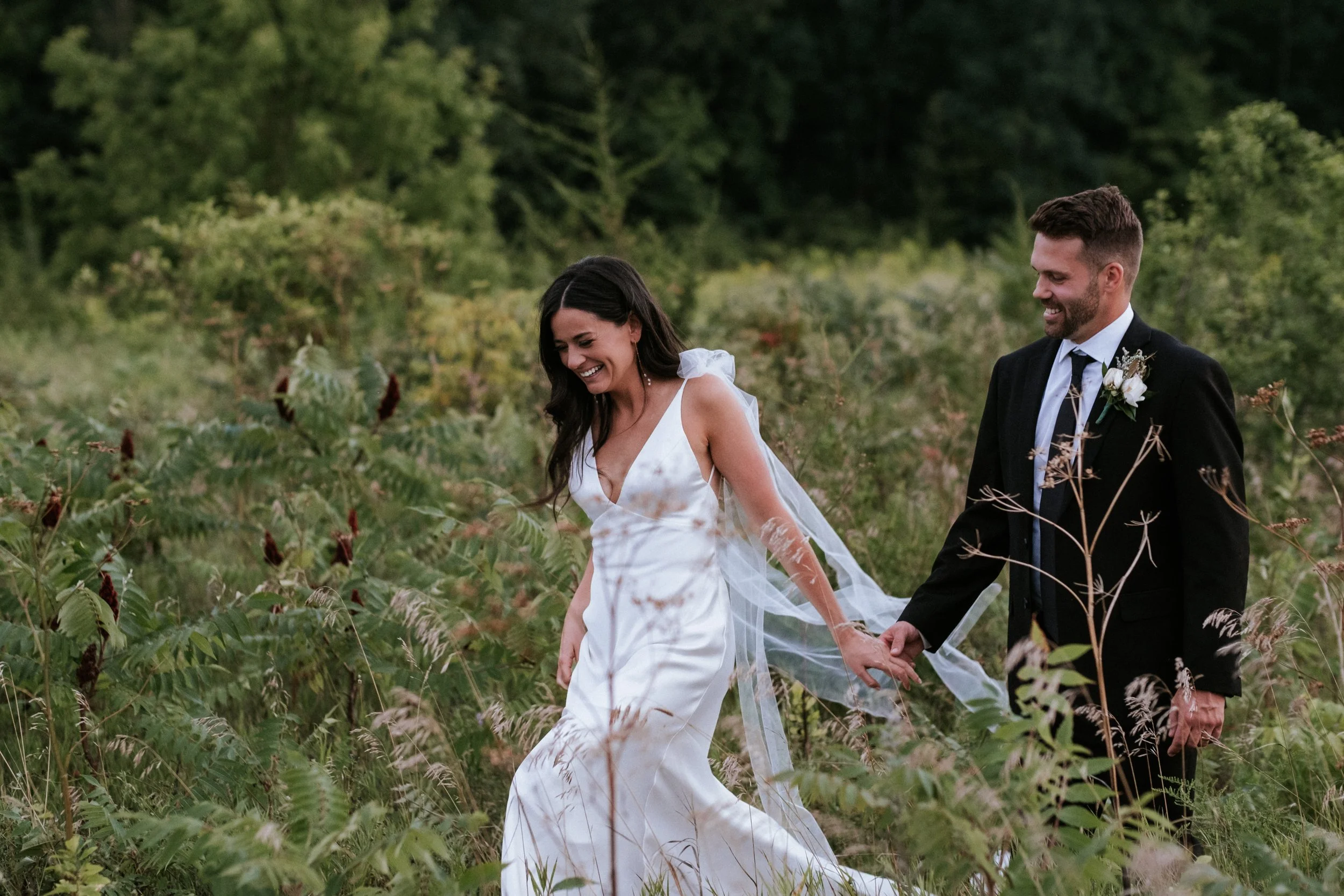 A bride and groom holding hands in a natural outdoor setting, smiling and looking joyful.