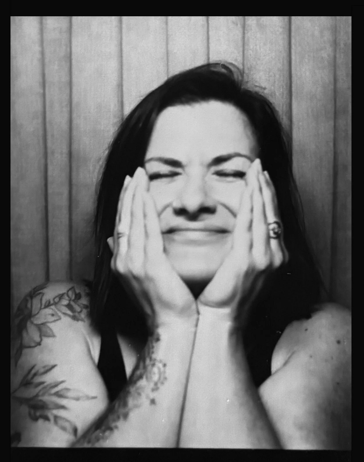 Black and white photo of a woman with dark hair, smiling with her eyes closed, holding her face with her hands, wearing rings and a bracelet, with a tattoo on her left arm, in front of a wooden panel background.