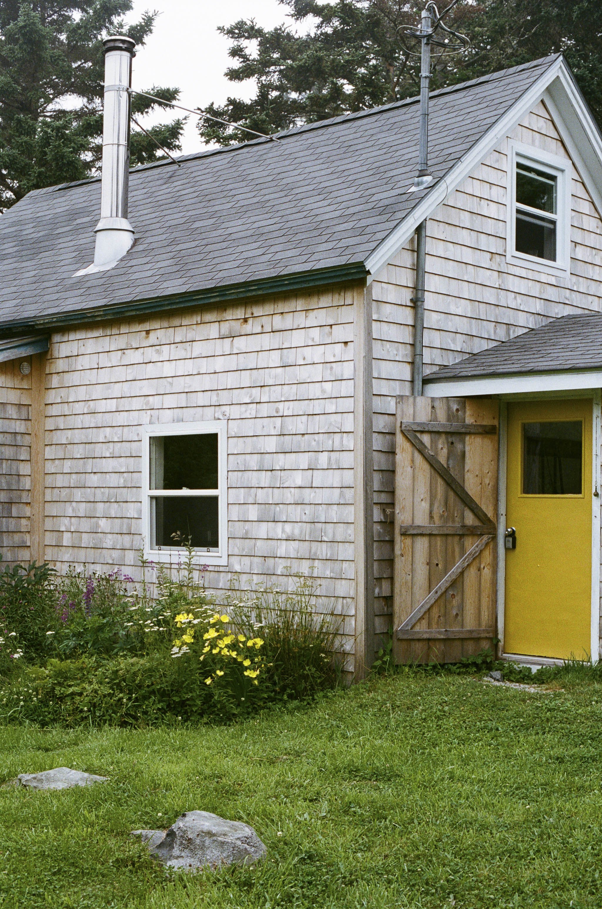 A two-story house with weathered wood siding, a yellow door, two double-hung windows, a gray shingled roof, a metal chimney, and a wooden fence gate, surrounded by green grass and flowering bushes.