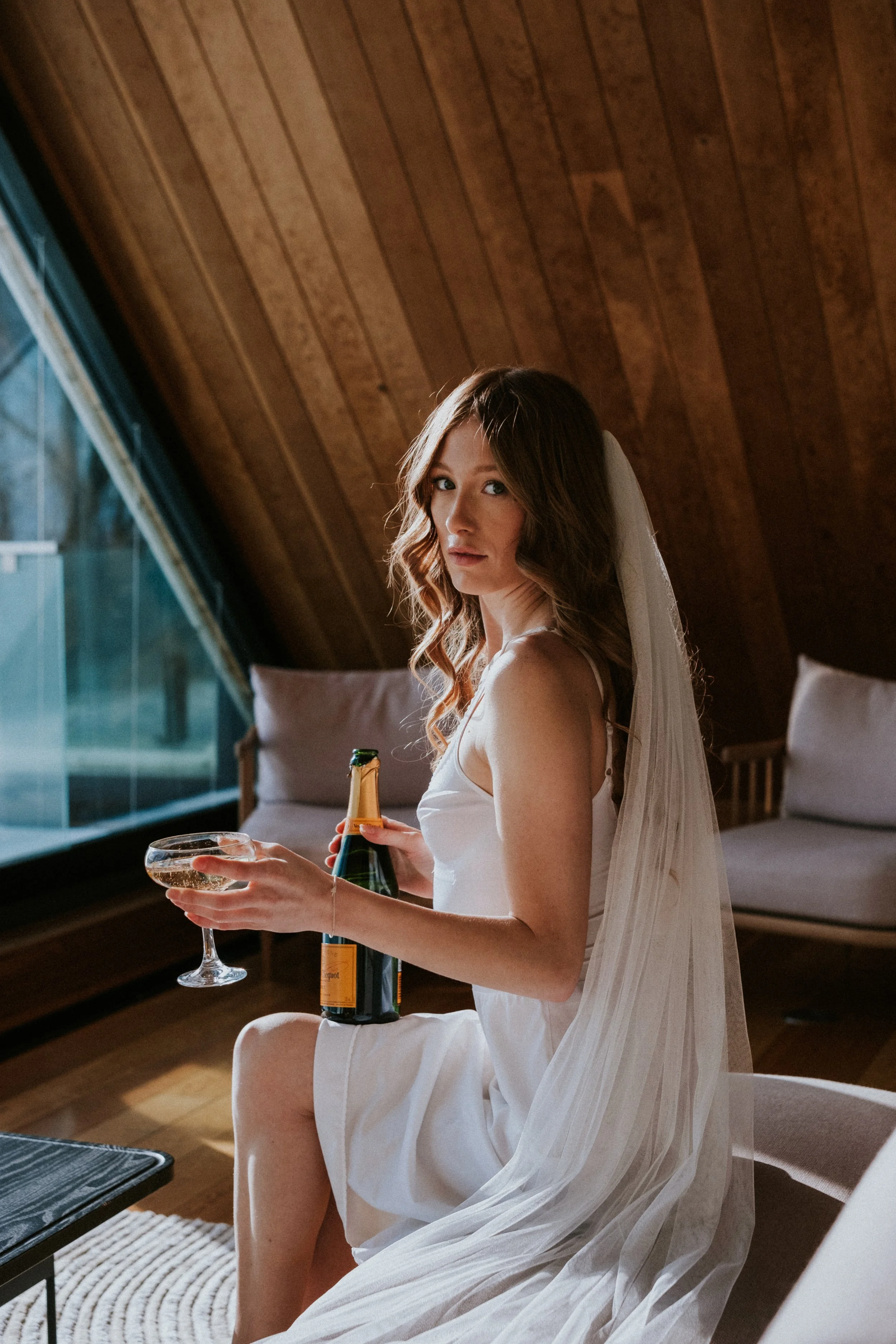 A young woman sitting indoors in a wooden-paneled room, wearing a white dress and veil, holding a champagne bottle and glass.