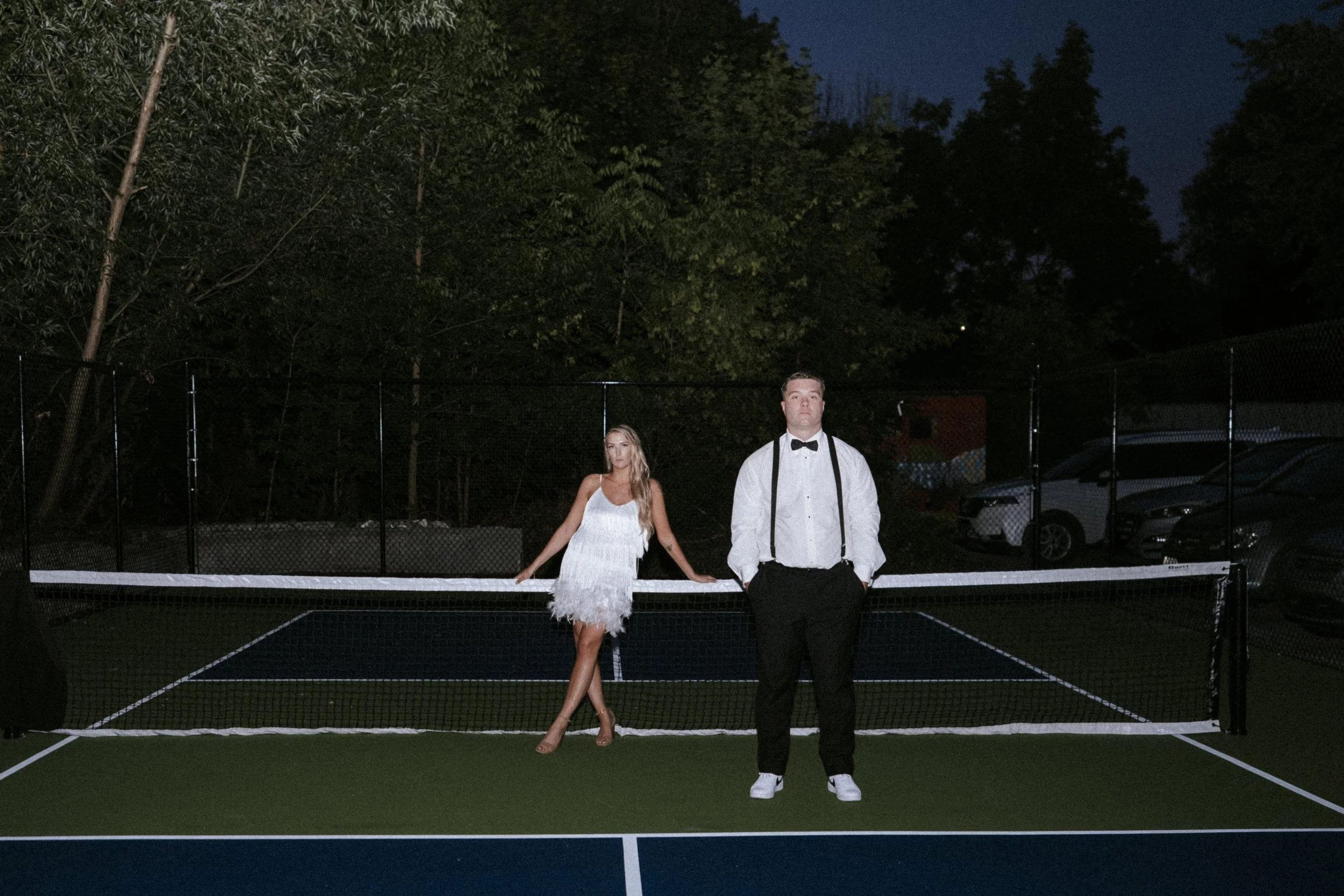 A man and woman in formal attire standing on a dark outdoor tennis court at night, with the woman leaning on the net and the man standing with his hands in his pockets.