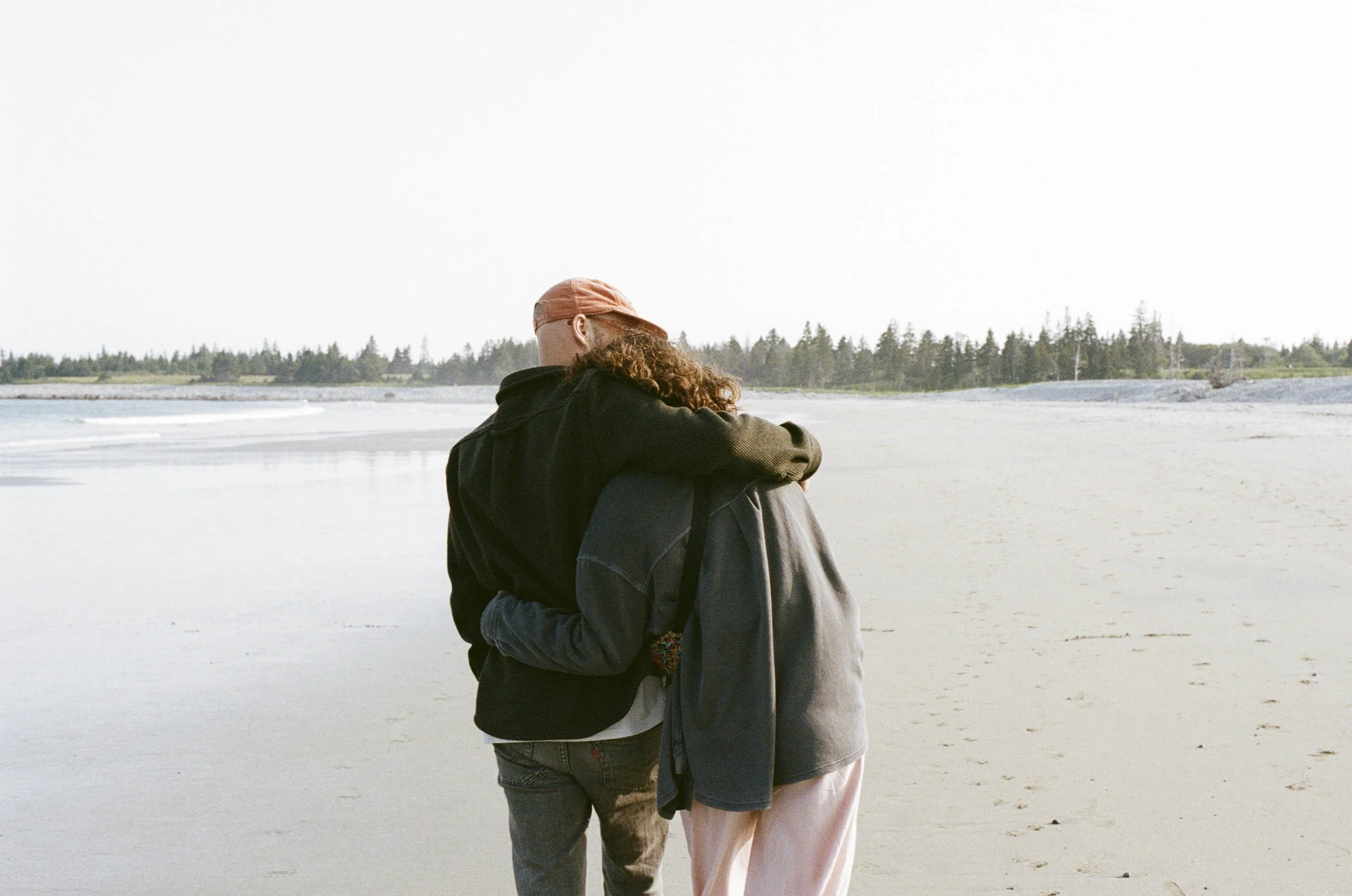 Two people hug on a beach with an ocean and trees in the background.