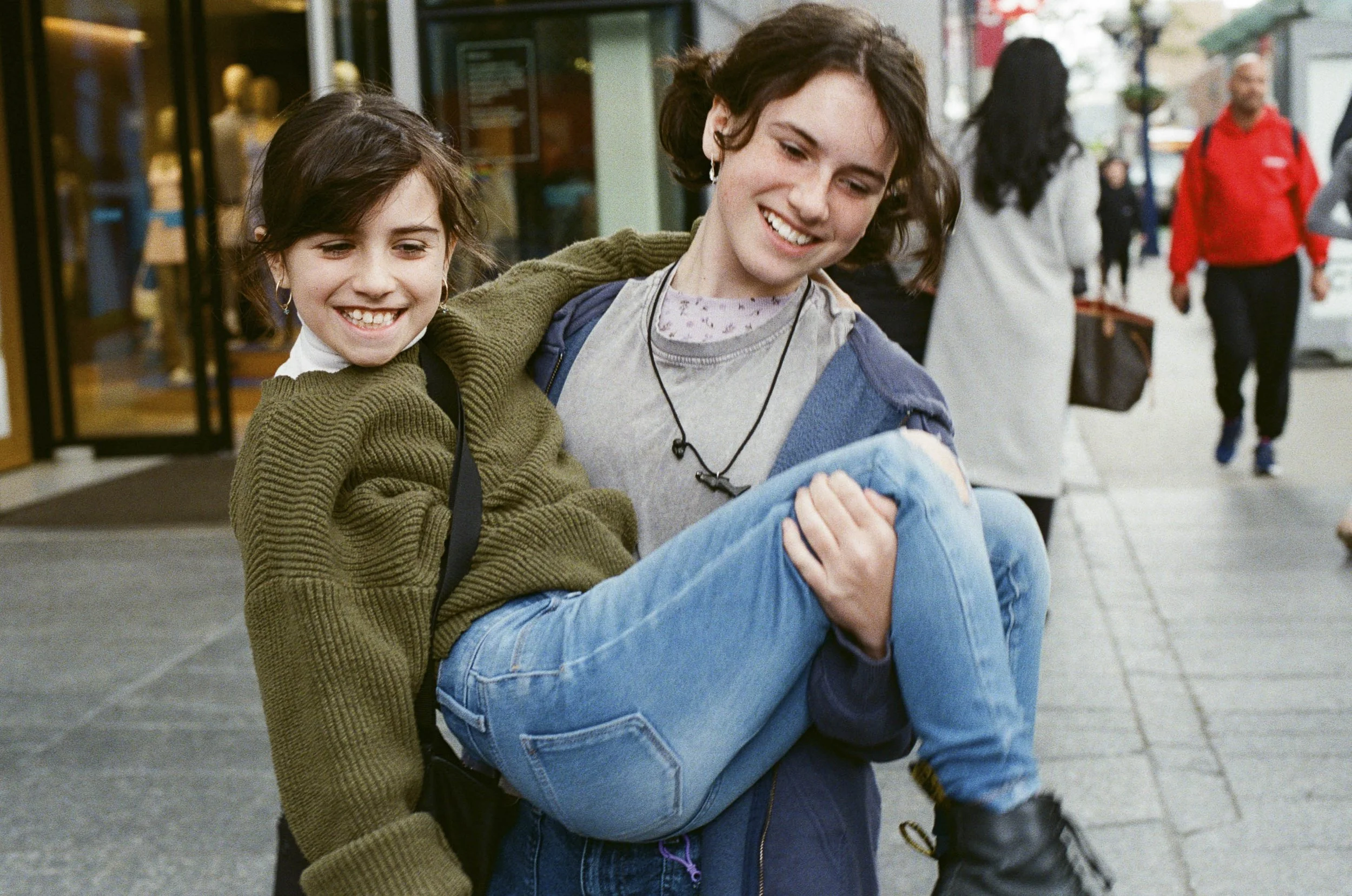 A young woman carrying a smiling girl on her lap on a city sidewalk, both looking down at something, with other pedestrians walking in the background.