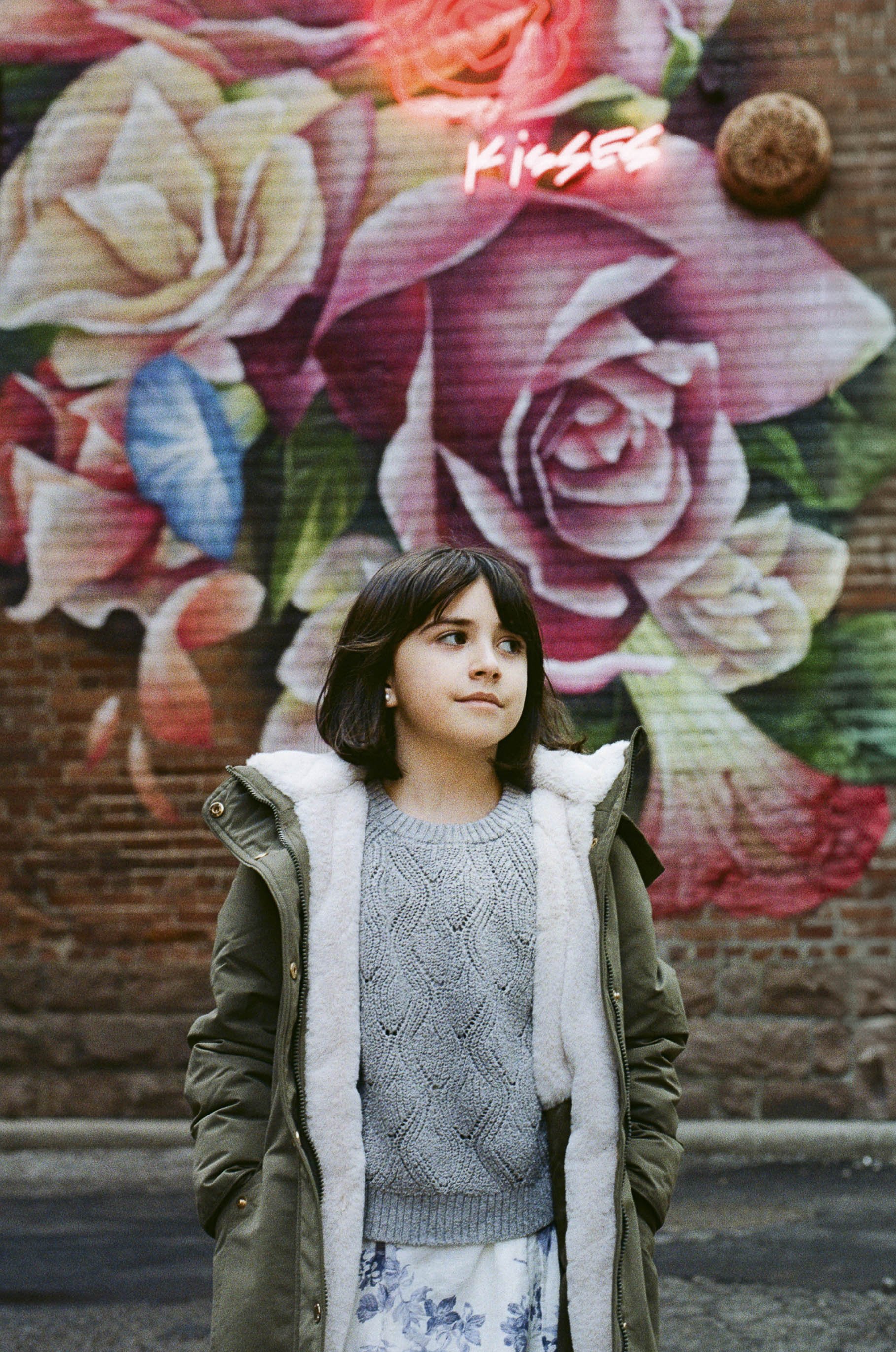 A young woman with dark hair in a green jacket and gray sweater standing in front of a large, colorful floral street art mural on a brick wall.