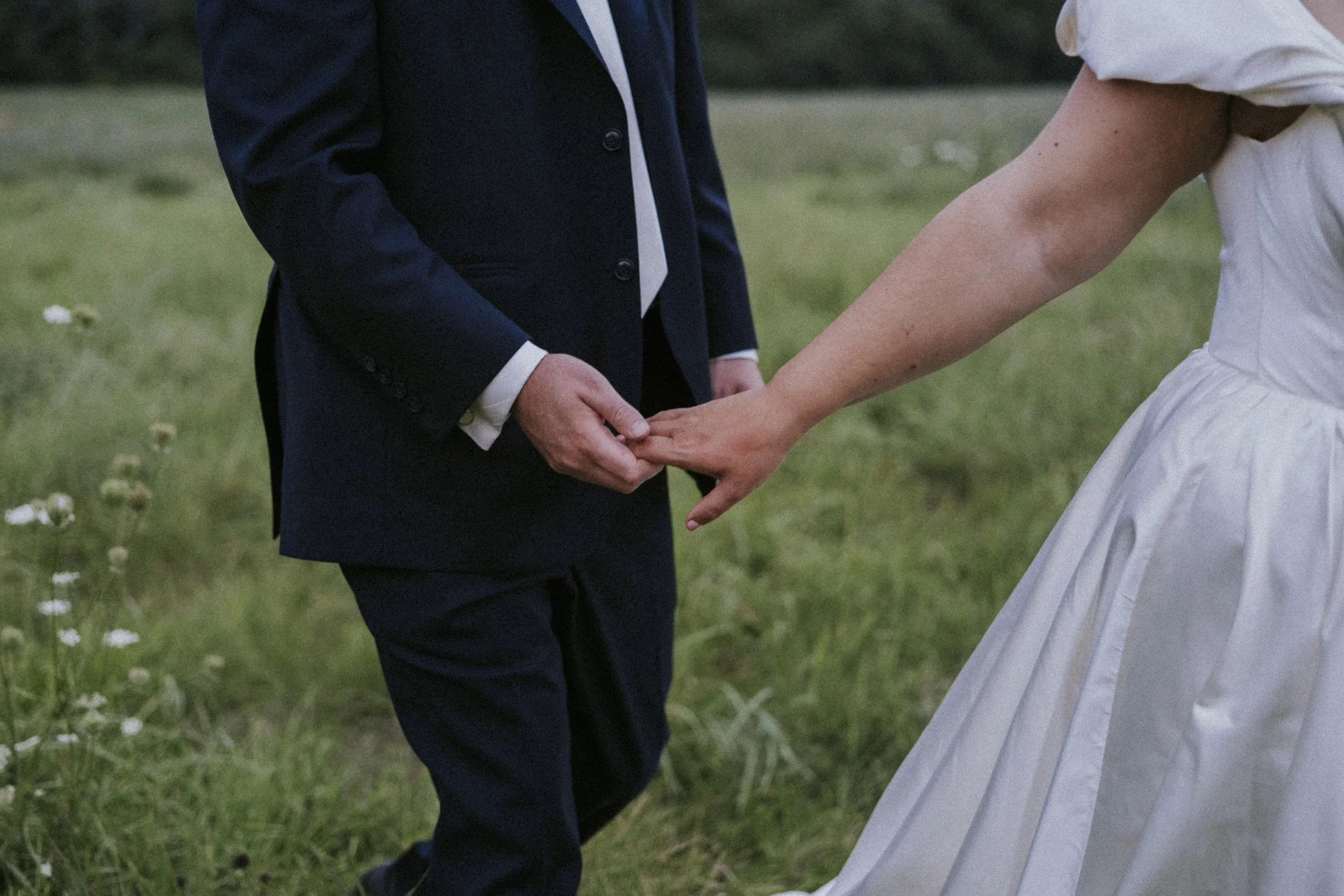 A person in a dark suit holding hands with a person in a white dress, outdoor setting with grass and small flowers.